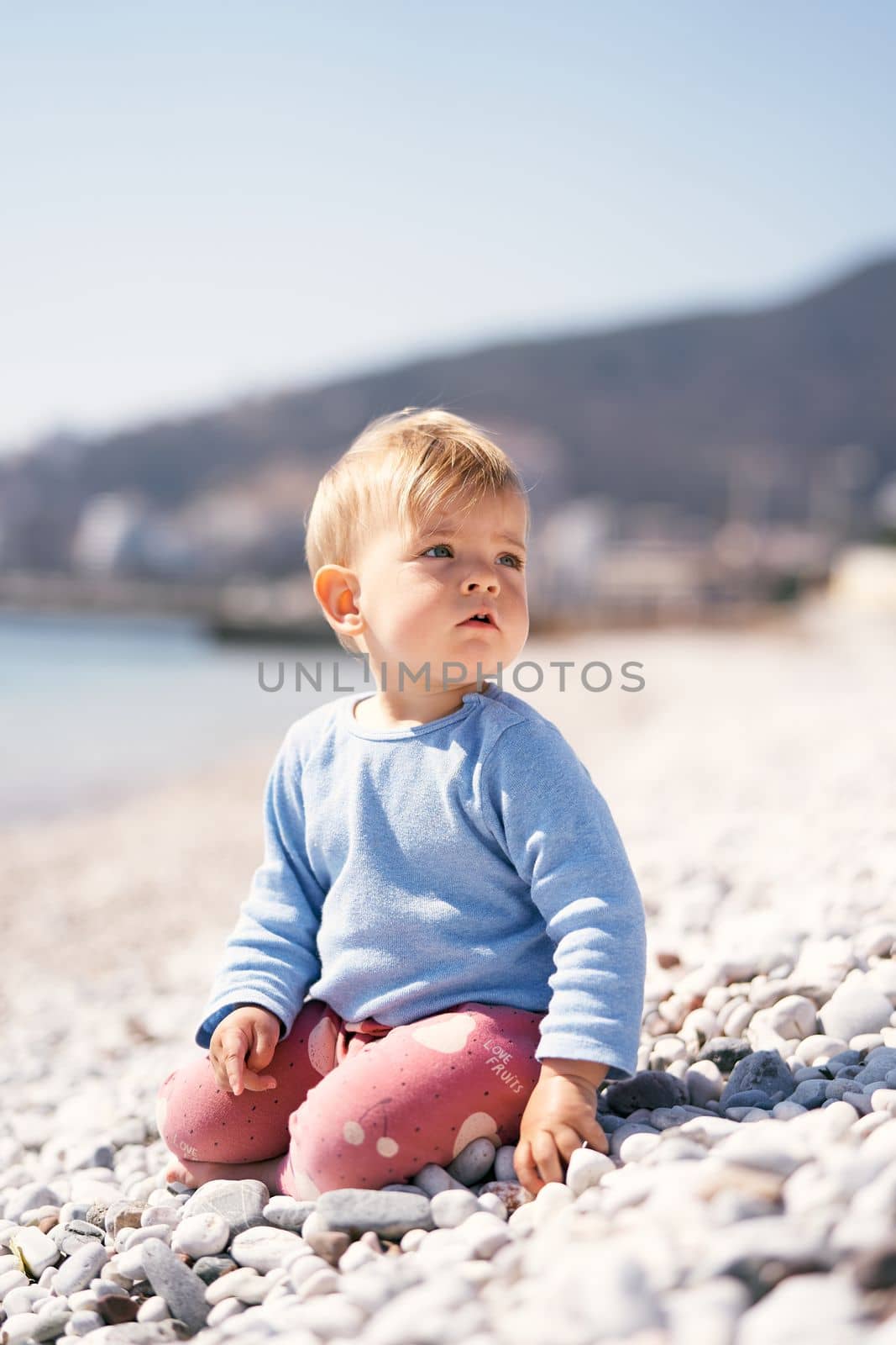 Pensive baby sits, turning to the right, on a pebble beach by the sea against a background of mountains by Nadtochiy