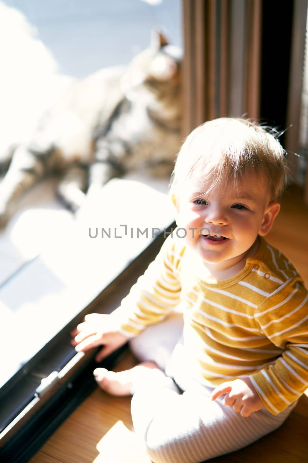 Laughing toddler sits on the floor near the open door to the courtyard. Close-up by Nadtochiy