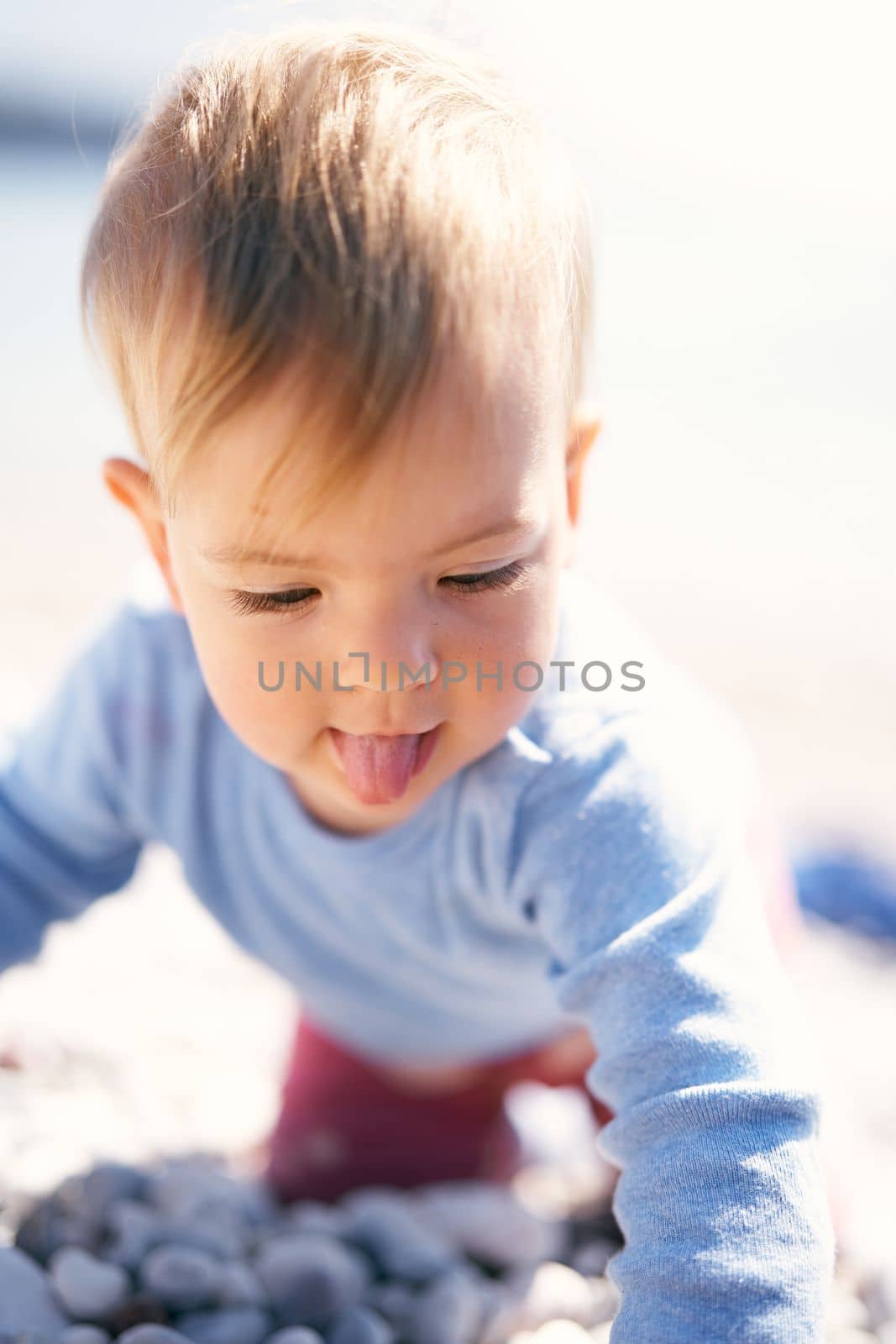 Cute baby, sticking out his tongue, sits on his knees on a pebble beach. Close-up by Nadtochiy