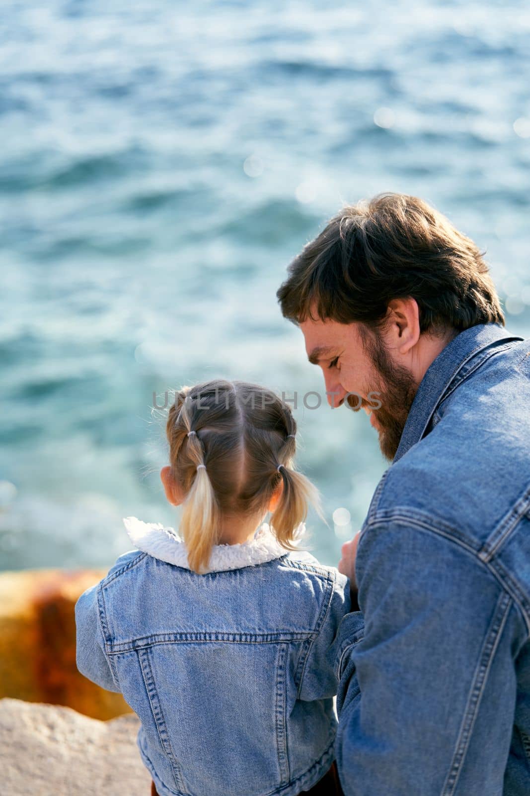 Smiling dad sits next to a little girl on a stone above the sea. Back view. Close-up. High quality photo