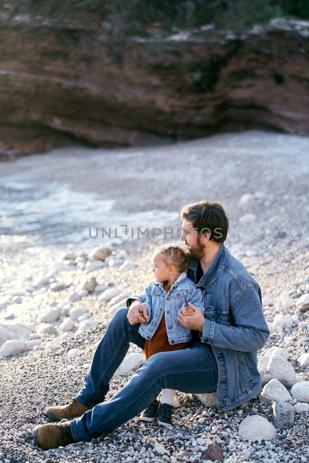 Dad sits on a stone and hugs a little girl on a pebble beach against the backdrop of rocks. Daughter and dad are looking at the sea. Side view by Nadtochiy