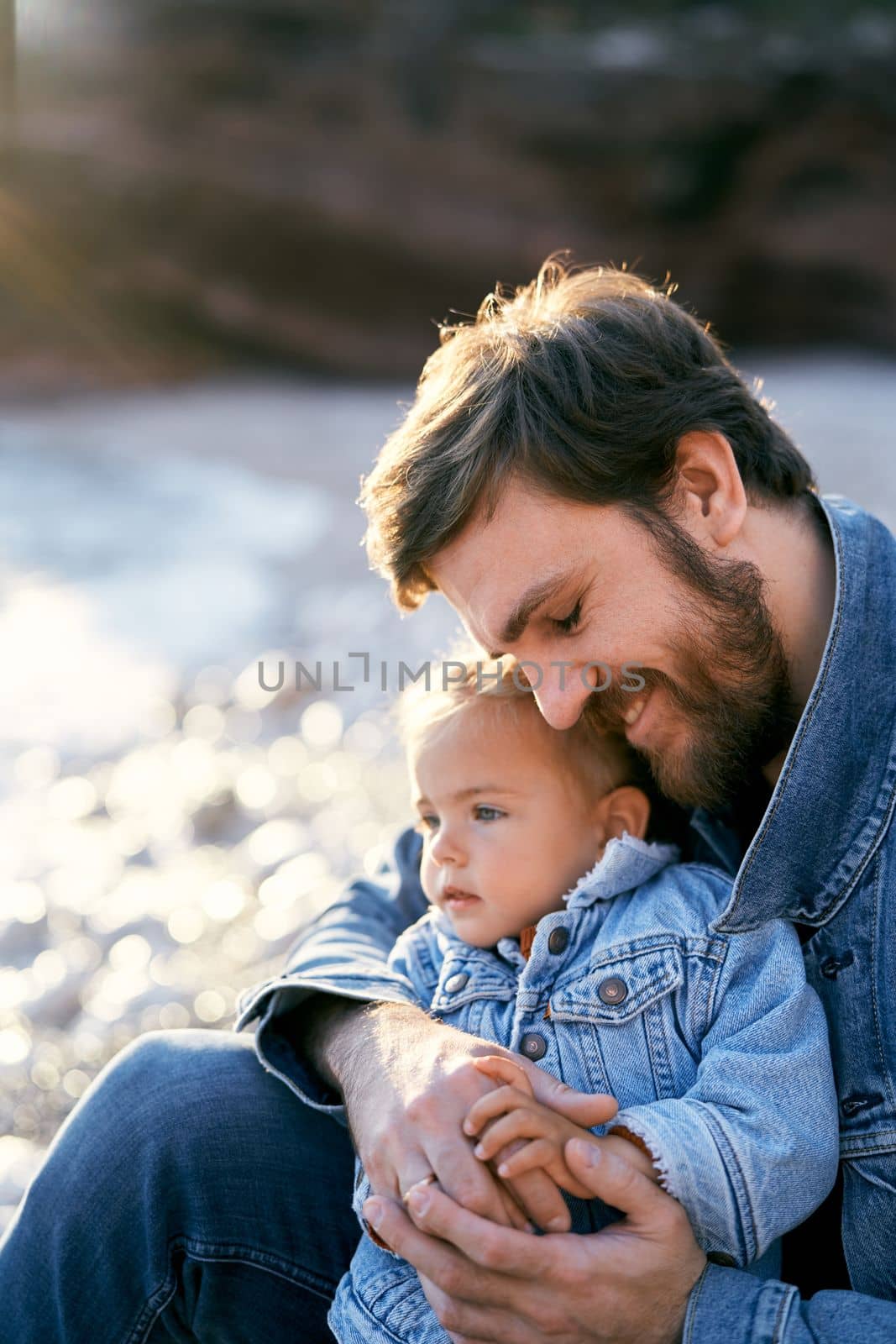 Smiling dad sits and hugs a little girl on a pebble beach. Close-up by Nadtochiy