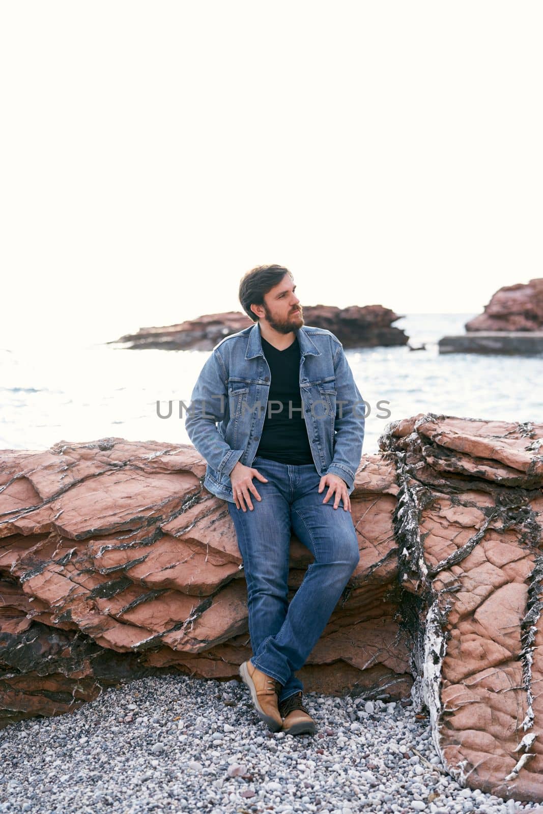 Pensive man in jeans and a denim jacket stands leaning against the stones near the sea and looks away. High quality photo