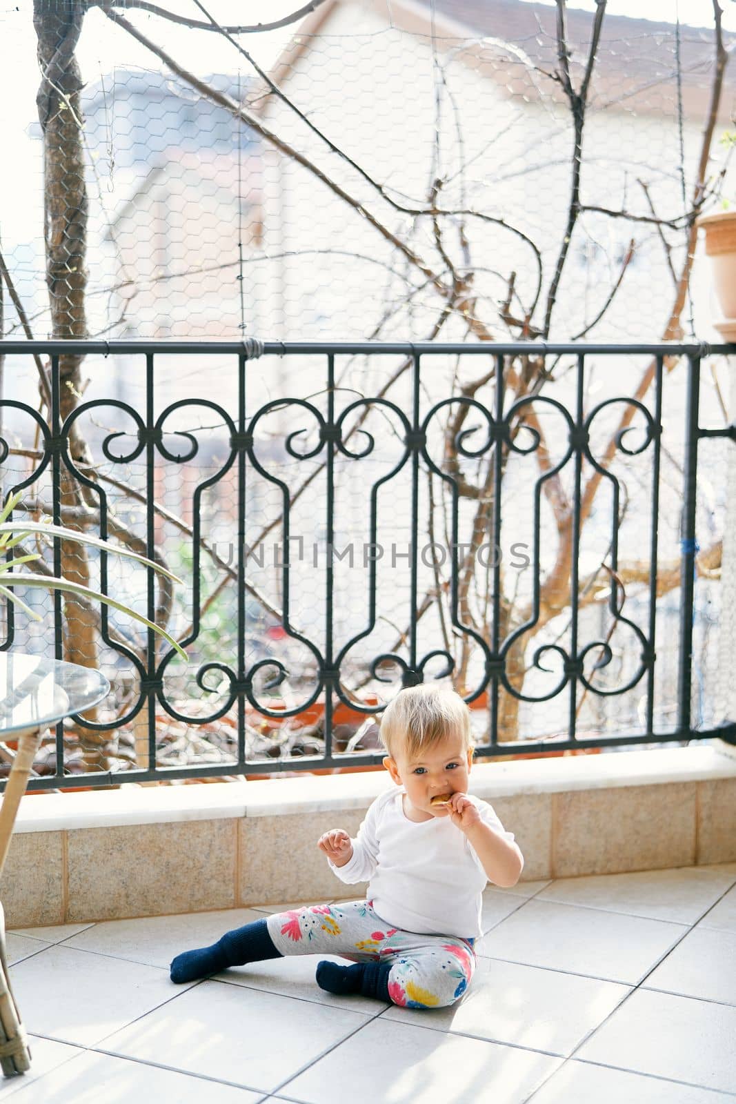 Small child in a white blouse and bright pants sits on the balcony near the table and gnaws cookies on the background of trees. High quality photo