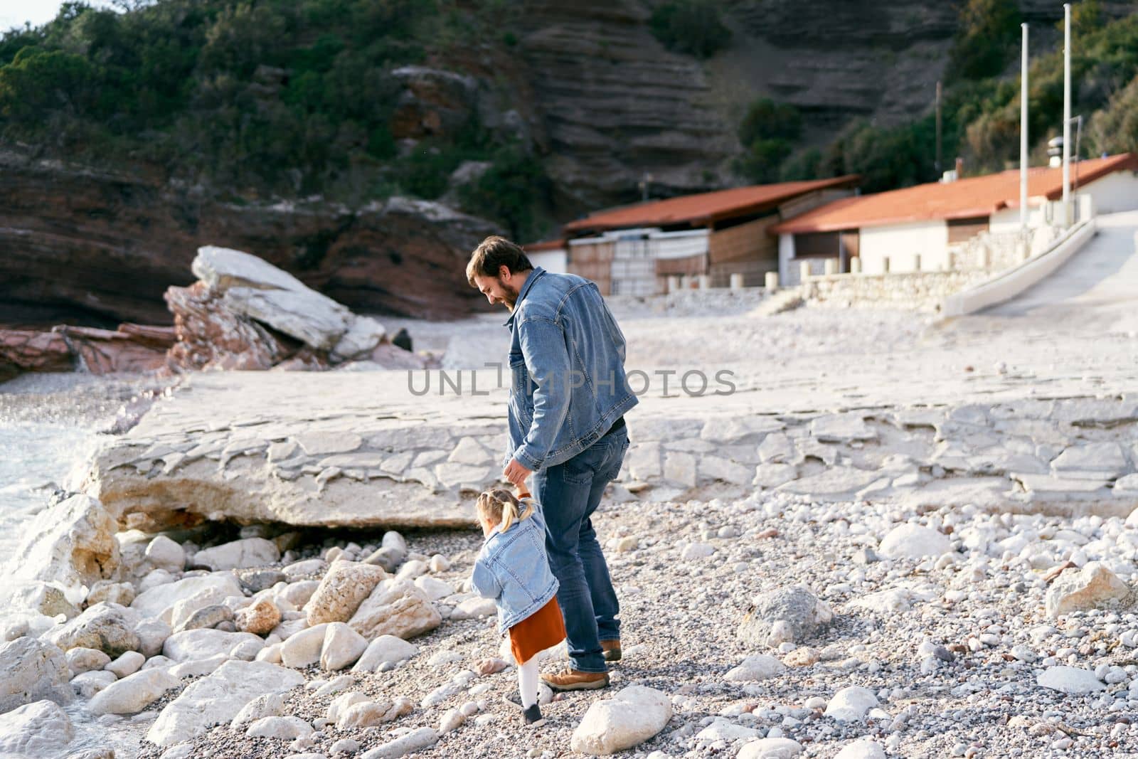 Dad leads a little girl by the hand along a pebble beach against the backdrop of buildings and rocky mountains. Back view. High quality photo