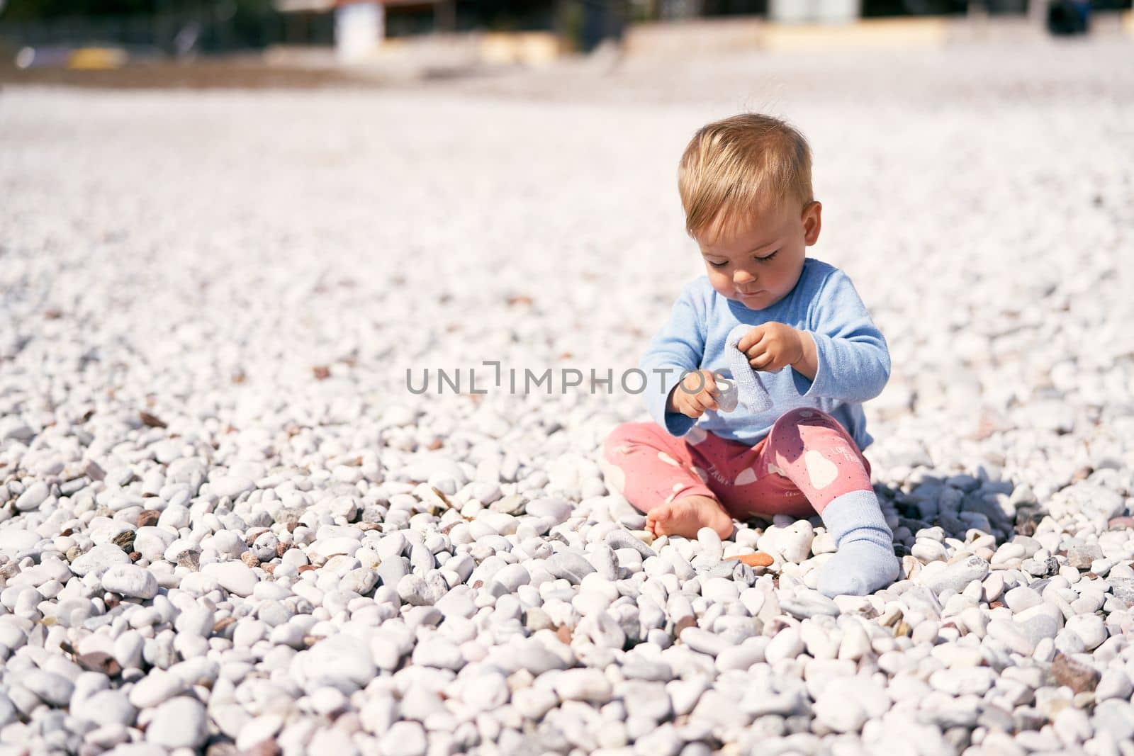 Cute baby in a blue blouse and red pants sits on a pebble beach, bowing his head and holding pebbles in his hands. High quality photo