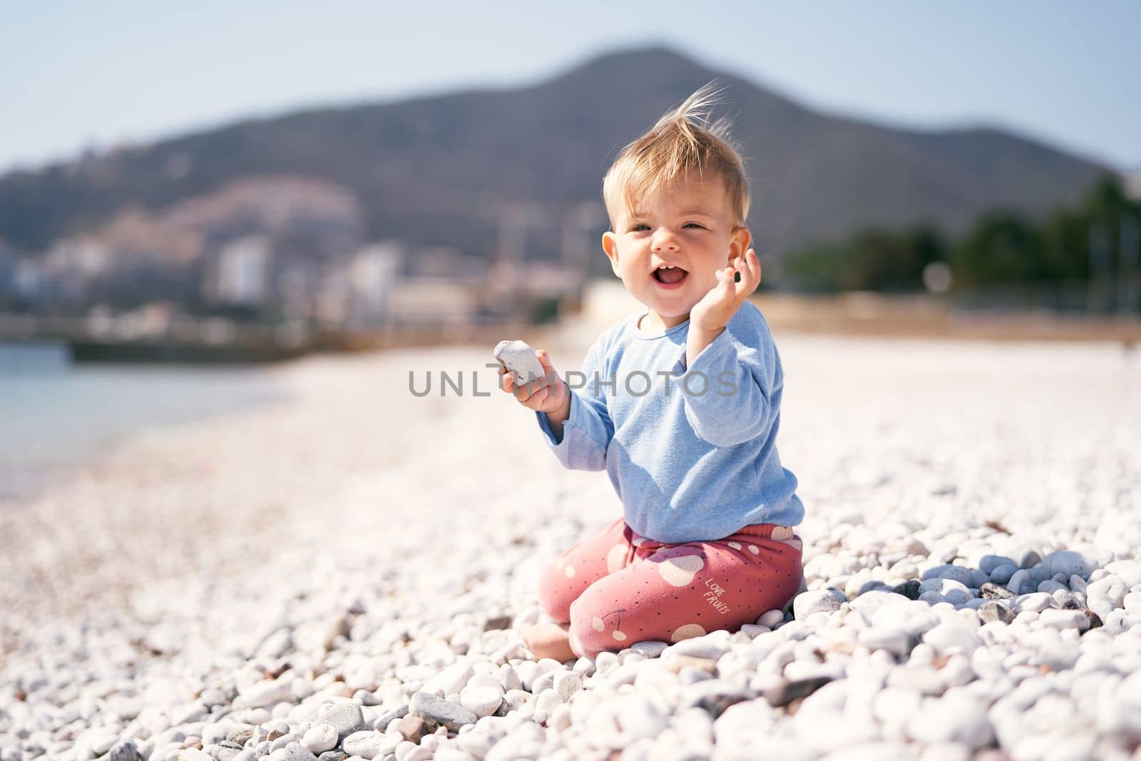 Laughing kid sits with a pebble in one hand and a raised second hand on a pebble beach against a background of mountains. High quality photo
