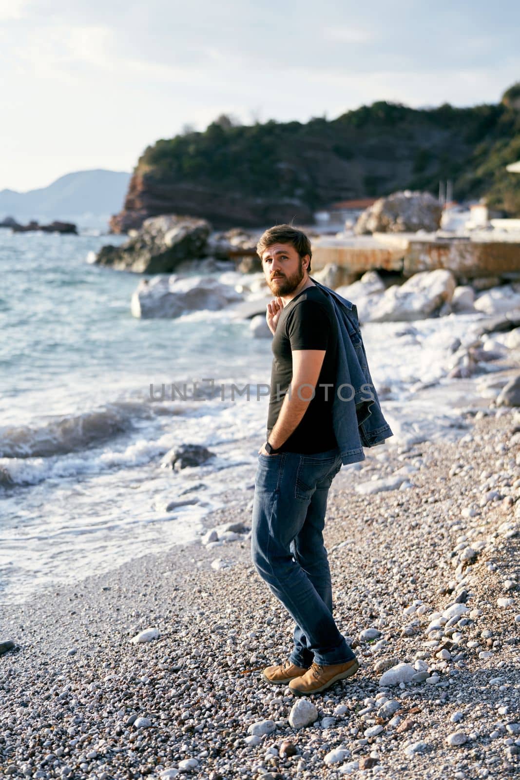 Pensive man stands on a pebble beach with a denim jacket over his shoulder against the background of the sea and rocks. High quality photo