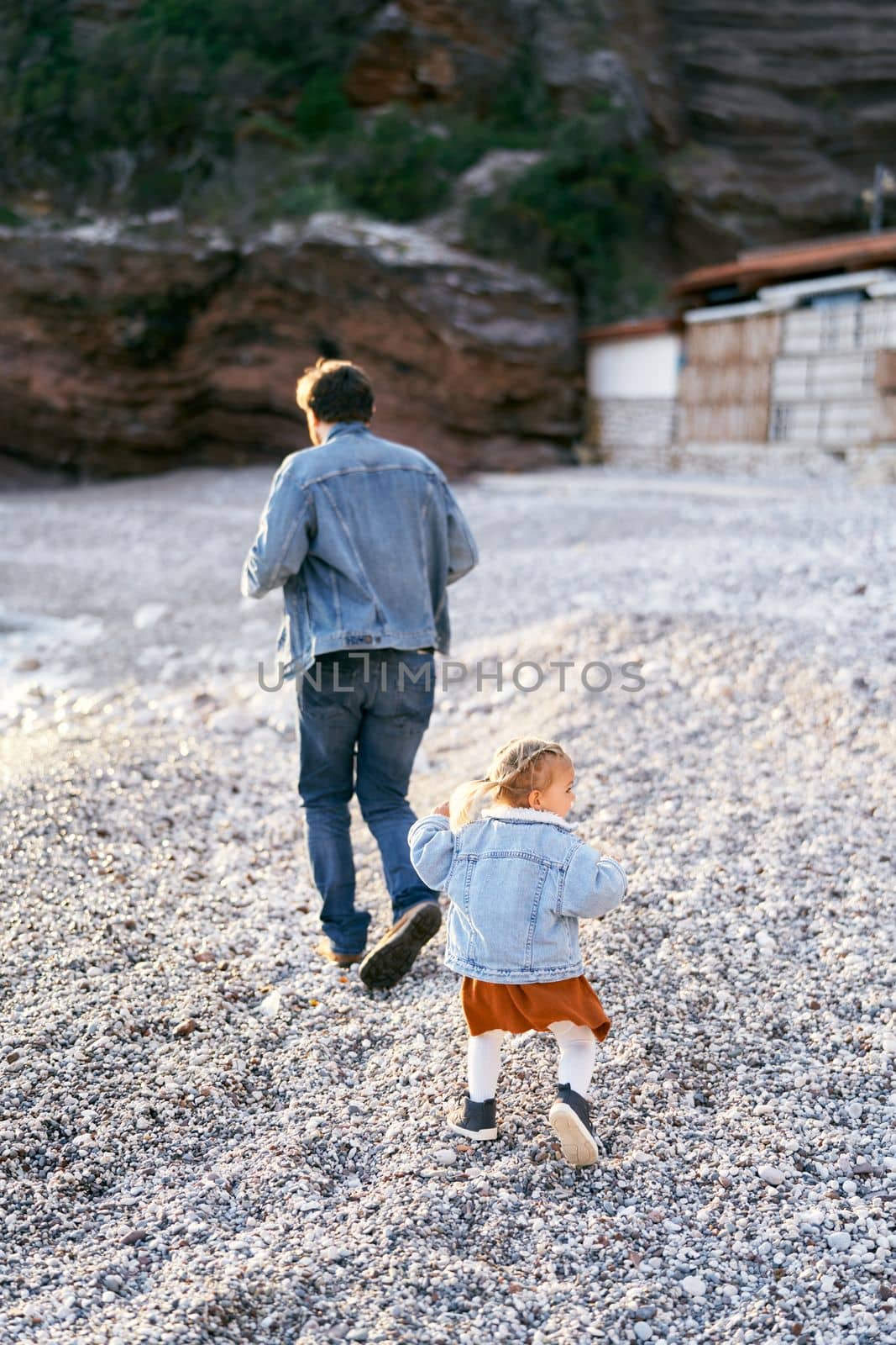Little girl runs after dad on the pebble beach against the backdrop of rocky mountains. Back view. High quality photo
