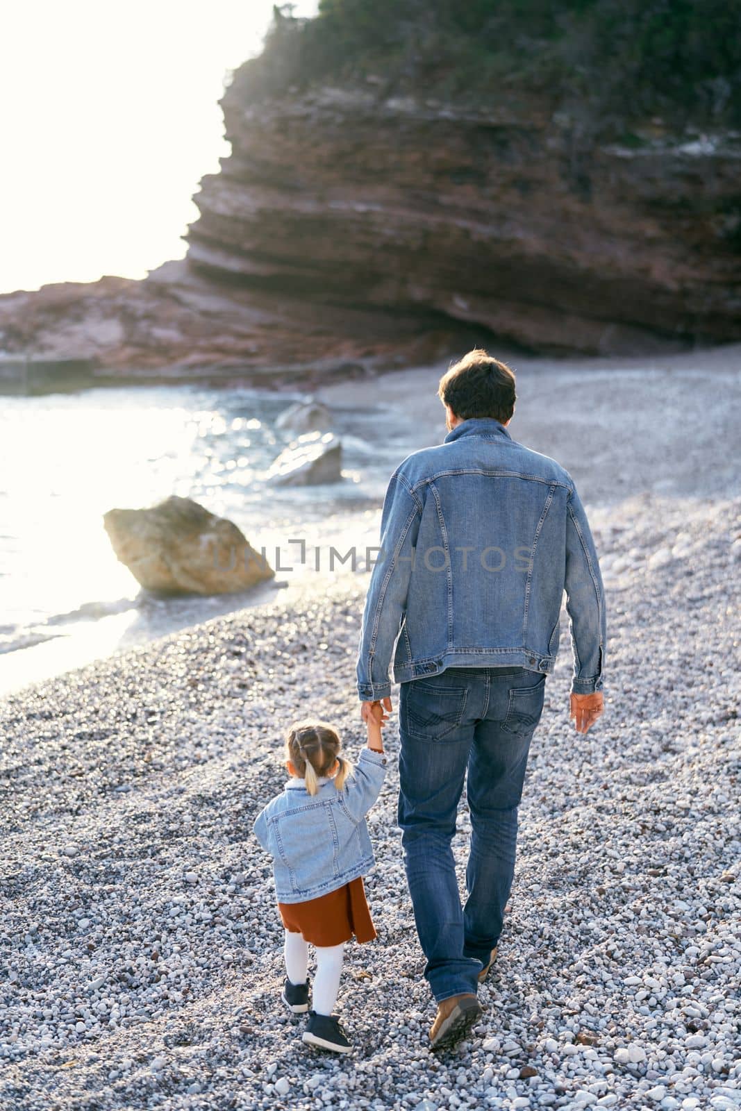 Dad leads a little daughter by the hand, walking along the pebble beach to rocky mountains and the sea. Back view. High quality photo
