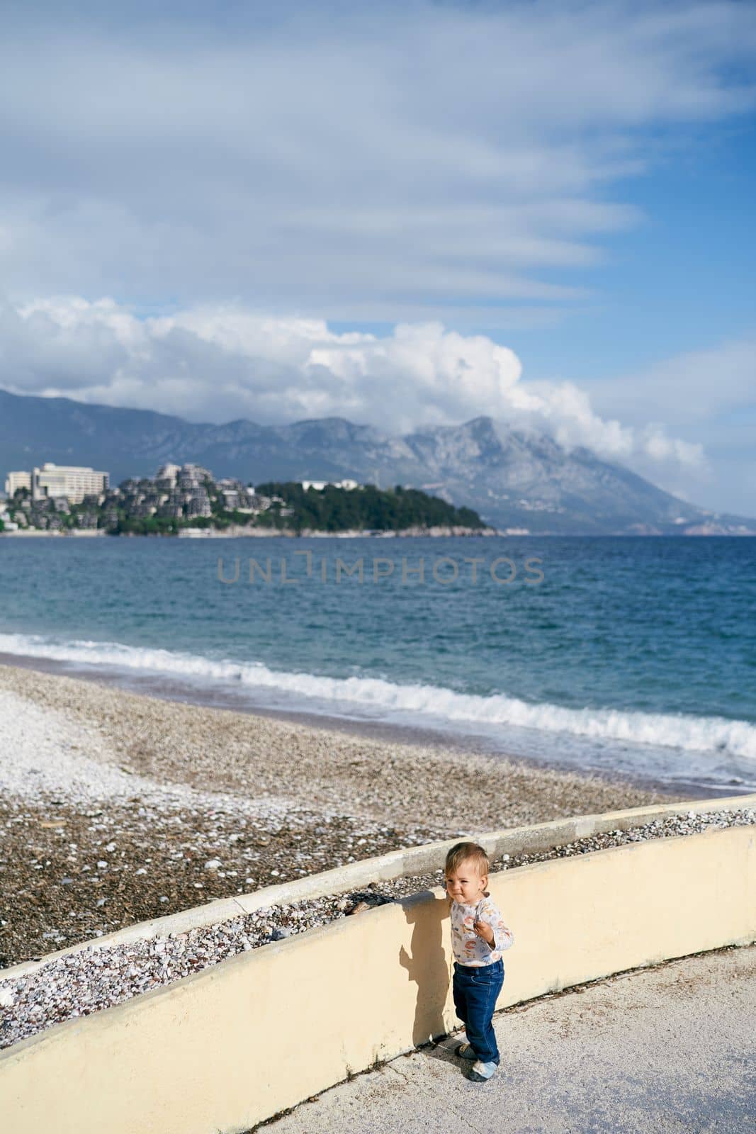 Kid stands on the beach near the concrete fence against the background of the sea. High quality photo