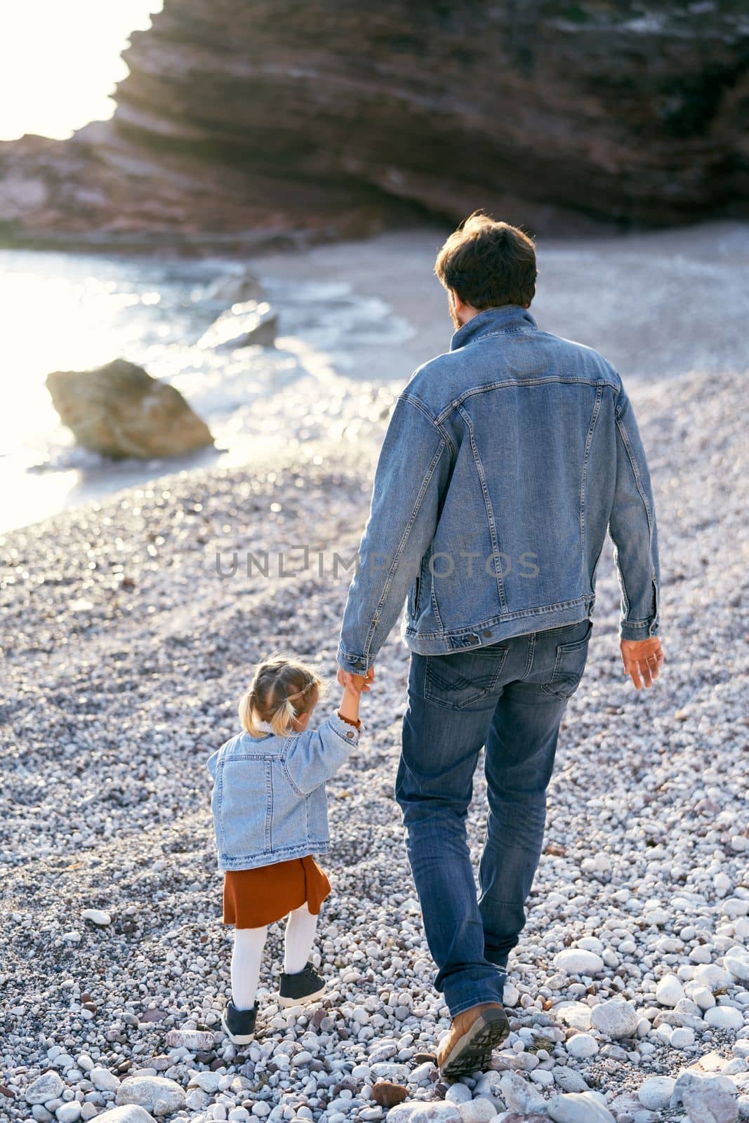 Dad leads a little girl by the hand, walking along the pebble beach against the backdrop of rocky mountains and the sea. Back view. High quality photo