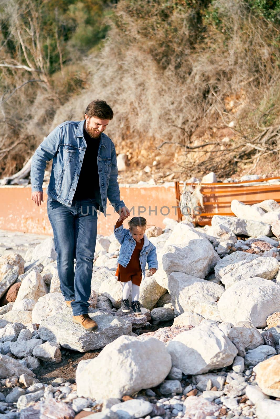 Smiling dad holds the hand of a little girl walking along the stones on a pebble beach against the backdrop of green mountains. High quality photo