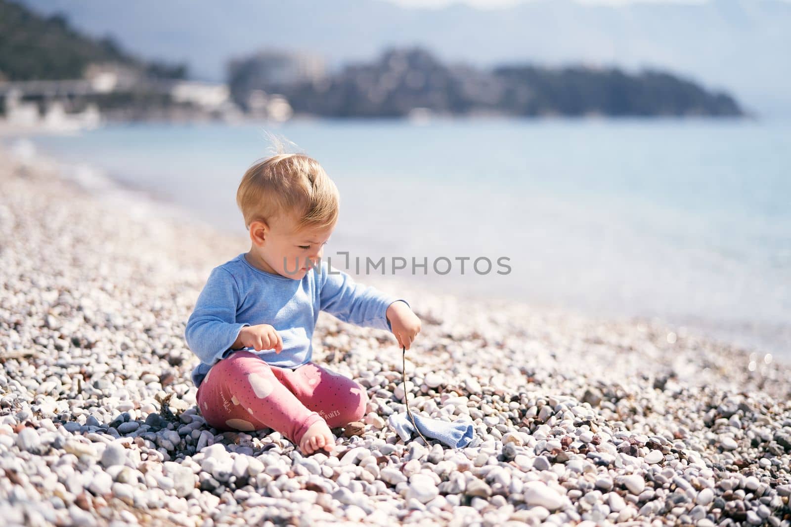 Little kid in a blue blouse and red pants sits on a pebble beach holding a stick in his hand. High quality photo