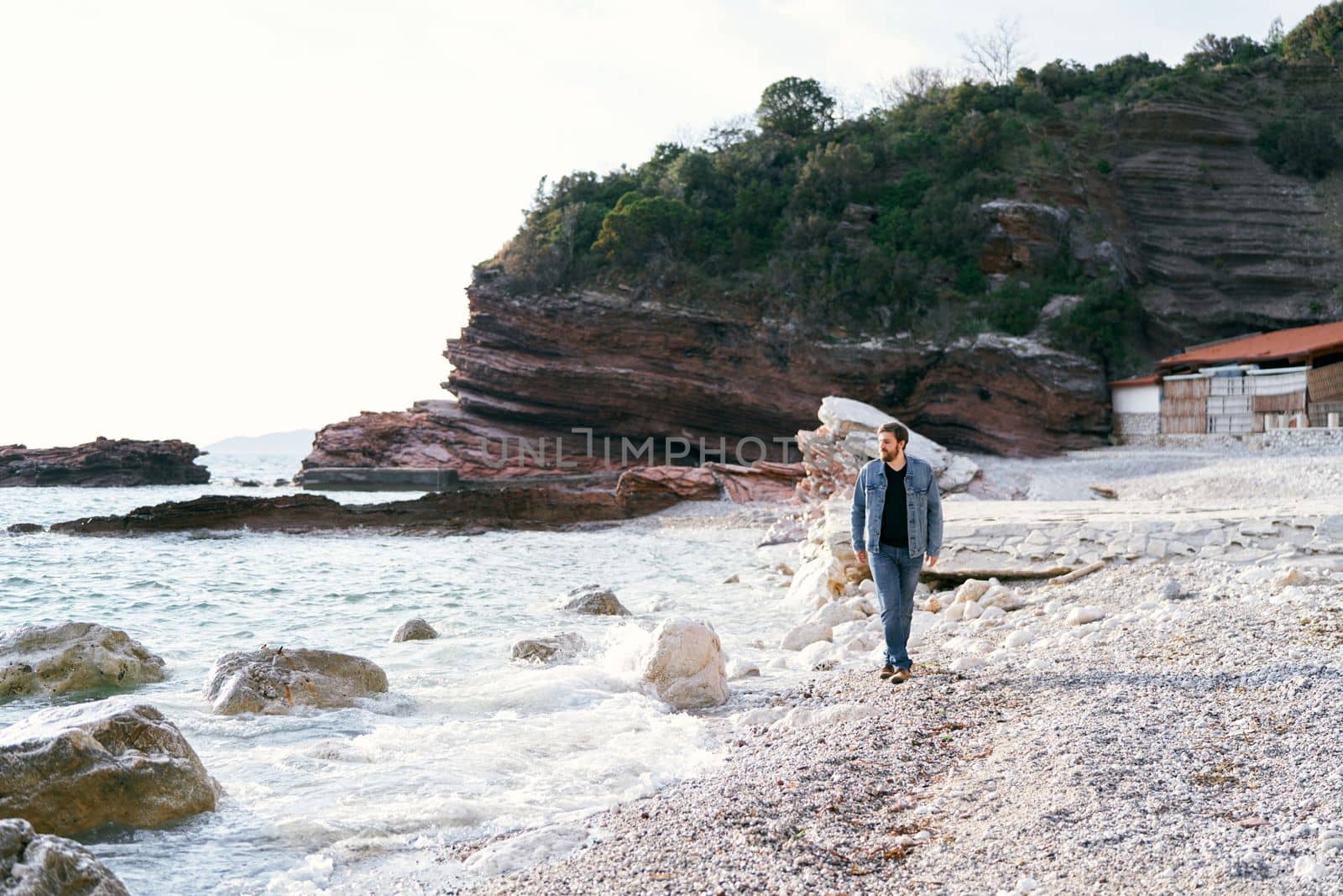 Pensive man in jeans and a denim jacket walks along a pebble beach looking at the water against a background of green rocks. High quality photo