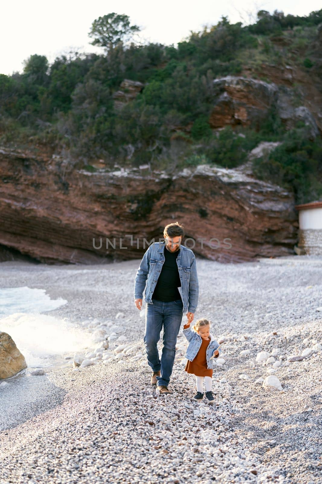 Smiling dad leads a little girl by the hand along a pebble beach against a background of green rocky mountains. High quality photo