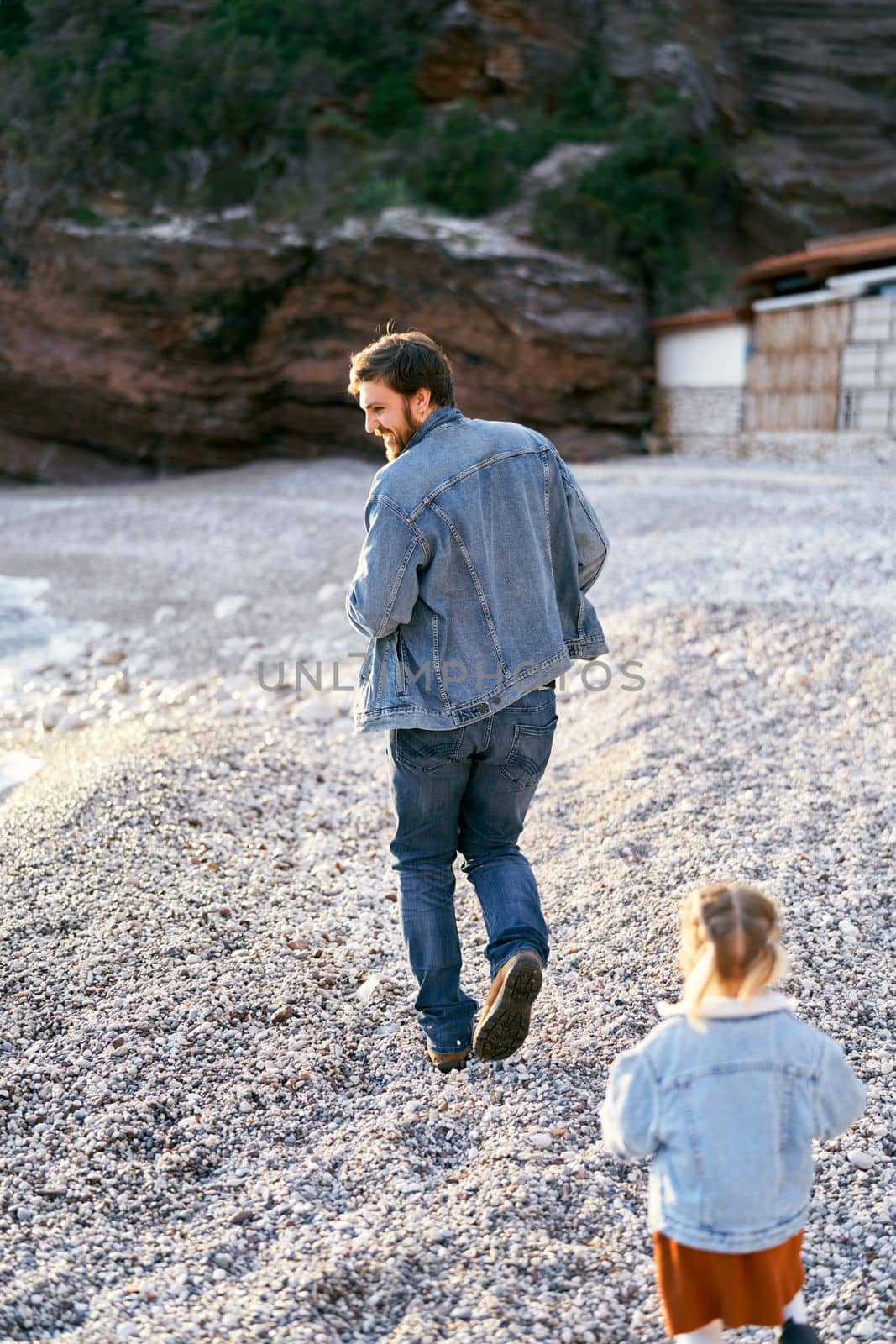 Little girl runs after smiling dad on the pebble beach against the backdrop of rocky mountains. High quality photo