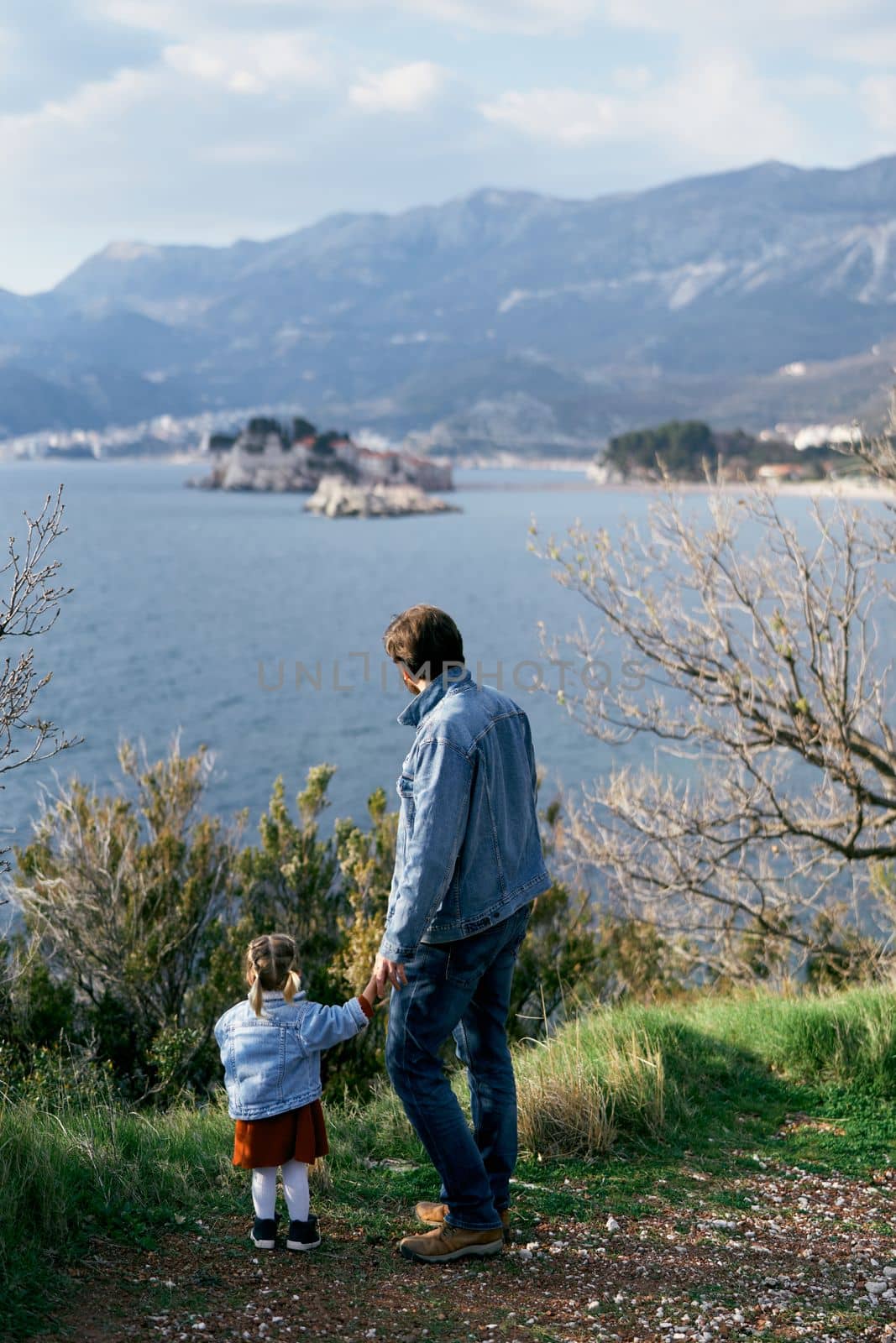Dad holds the hand of a little girl with a soft toy, standing on the coast against the background of the sea, mountains and Sveti Stefan island. Back view. High quality photo