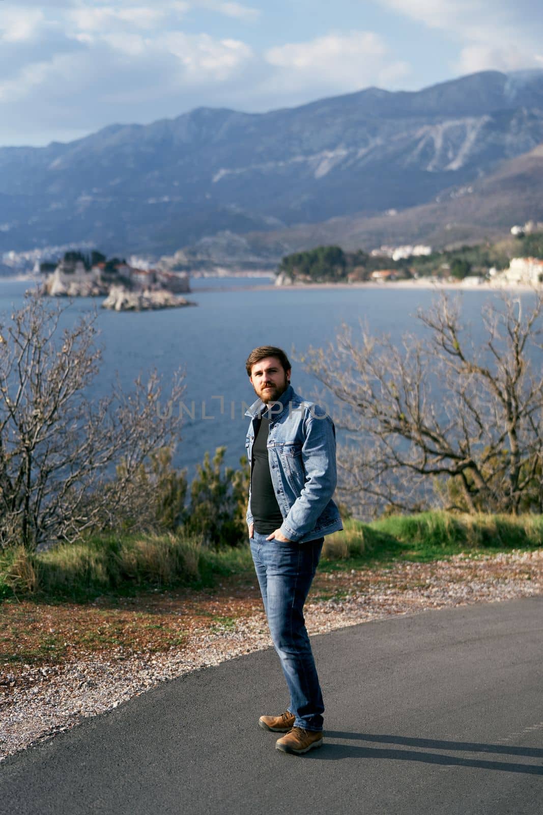 Man in a blue denim jacket and jeans stands holding his hands in his pockets on an asphalt road against the backdrop of the sea, mountains and the island of Sveti Stefan. High quality photo