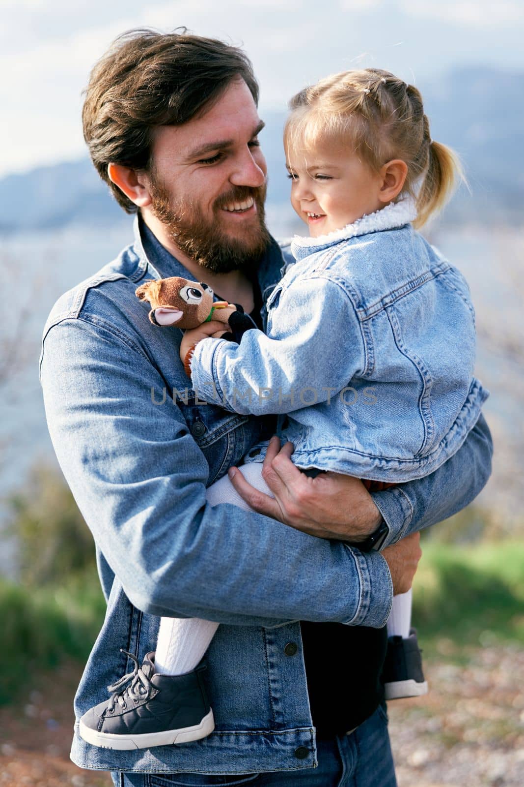 Smiling dad holds in his arms a smiling little girl with a soft toy standing on the background of the sea, mountains and the island of Sveti Stefan. Close-up. High quality photo