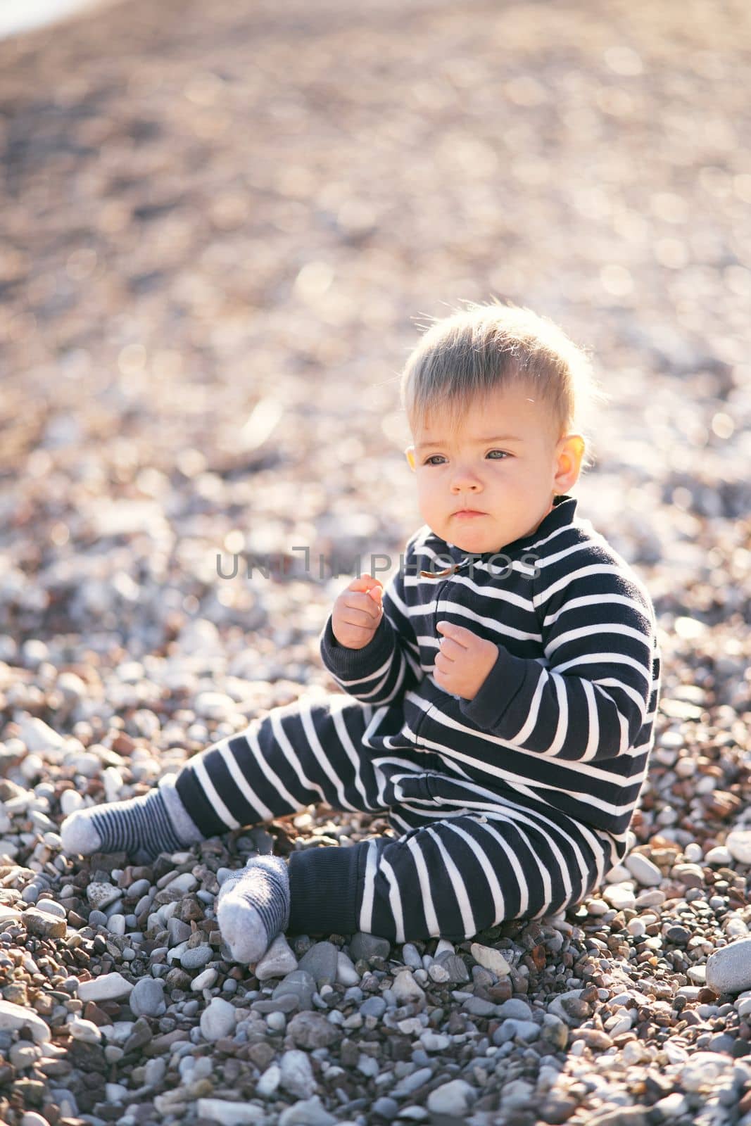 Serious toddler in striped overalls sits on a pebble beach, holding his hands in fists in front of him by Nadtochiy