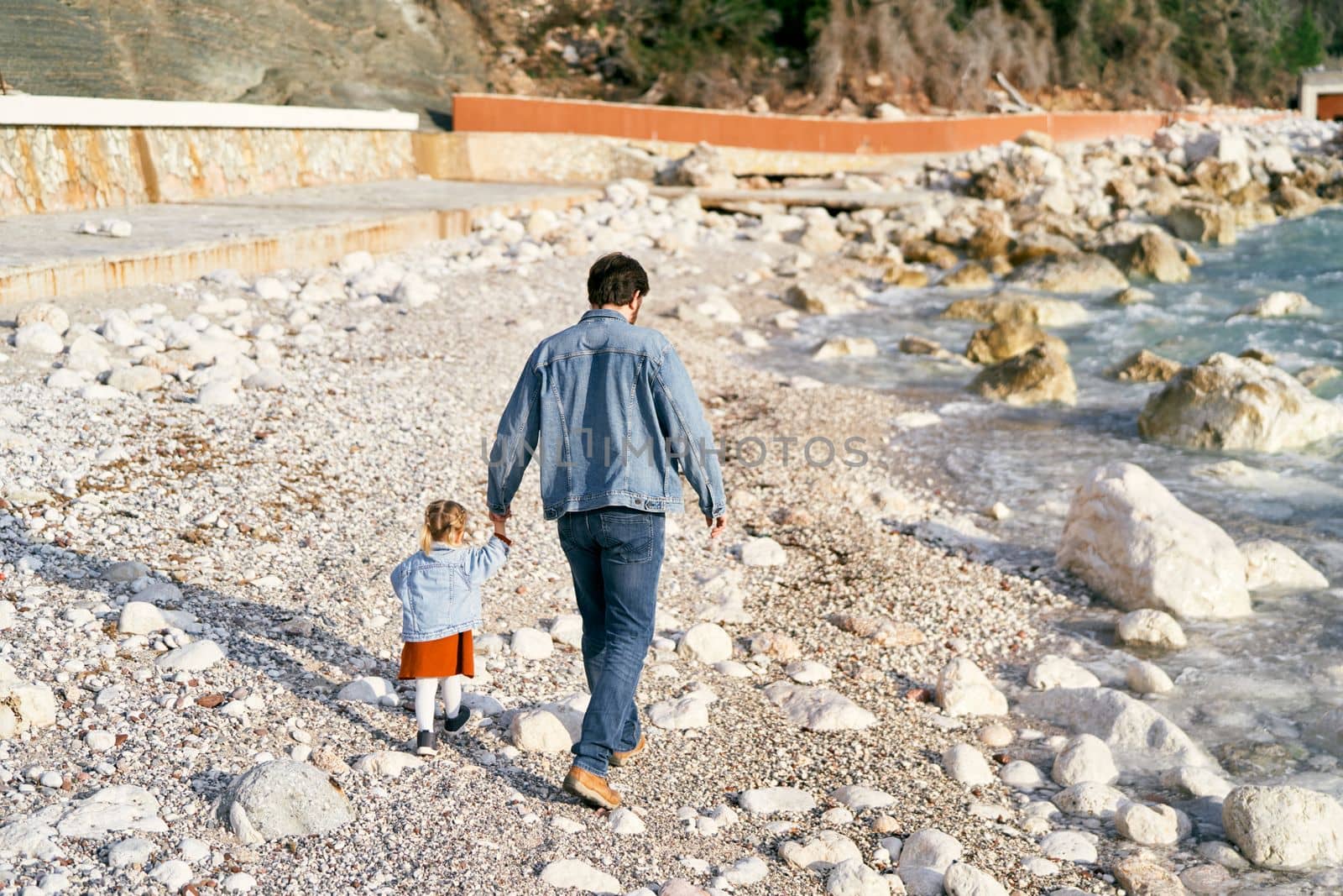 Dad in a denim jacket and jeans holds a little girl by the hand, walking with her along the rocky shore near the water. Back view. High quality photo
