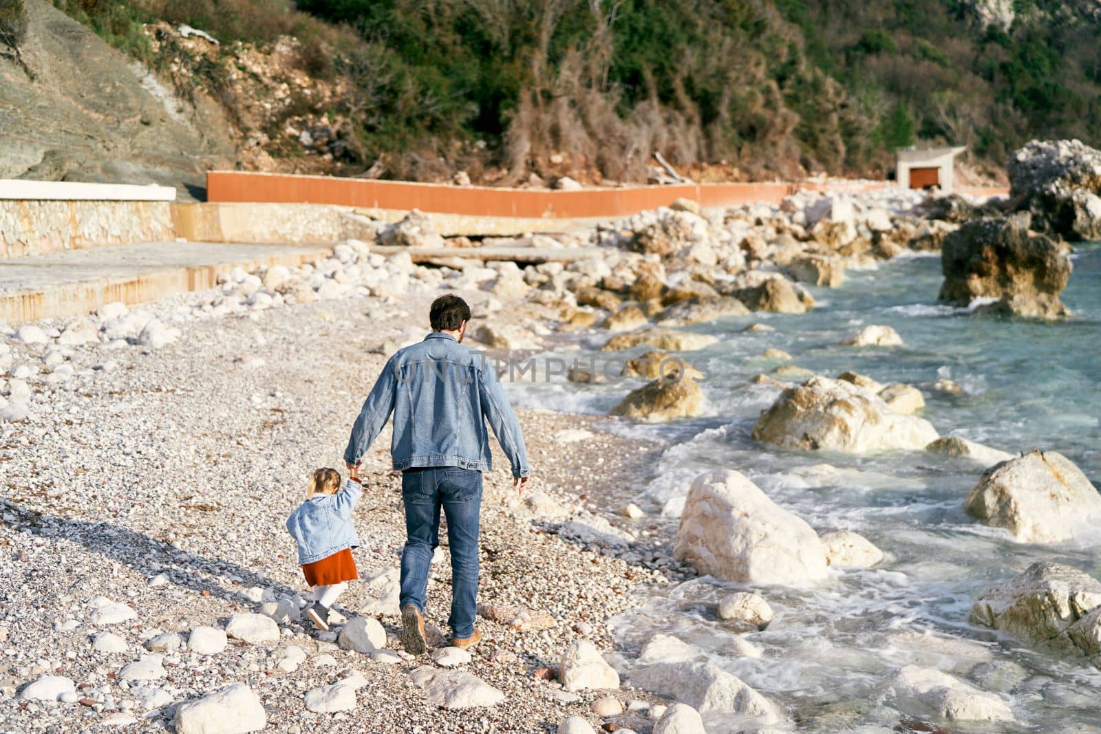 Dad in a denim jacket and jeans walks with a little girl holding her hand along a rocky pebble beach near the water. Back view. High quality photo