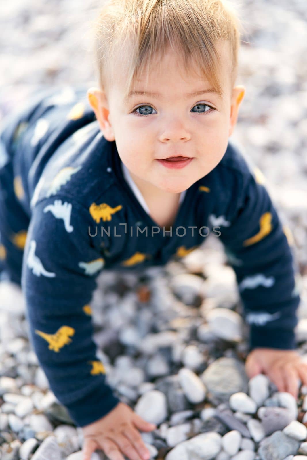 Smiling little baby in overalls sits on a pebble beach. Close-up. Portrait by Nadtochiy