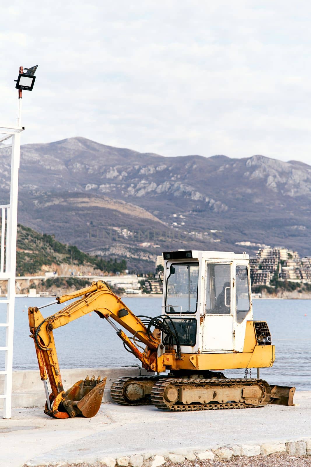 Yellow bulldozer stands on a stone coast against the background of the sea, houses on the other side and mountains by Nadtochiy