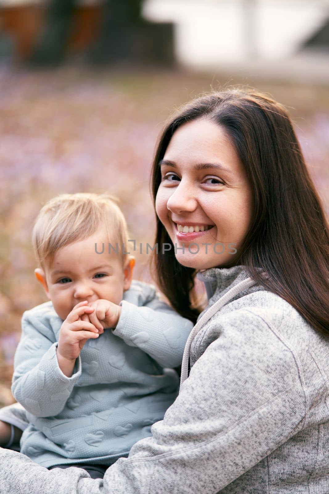 Smiling mom hugs toddler with hands in his mouth while sitting on foliage in the park. Close-up. Portrait. High quality photo