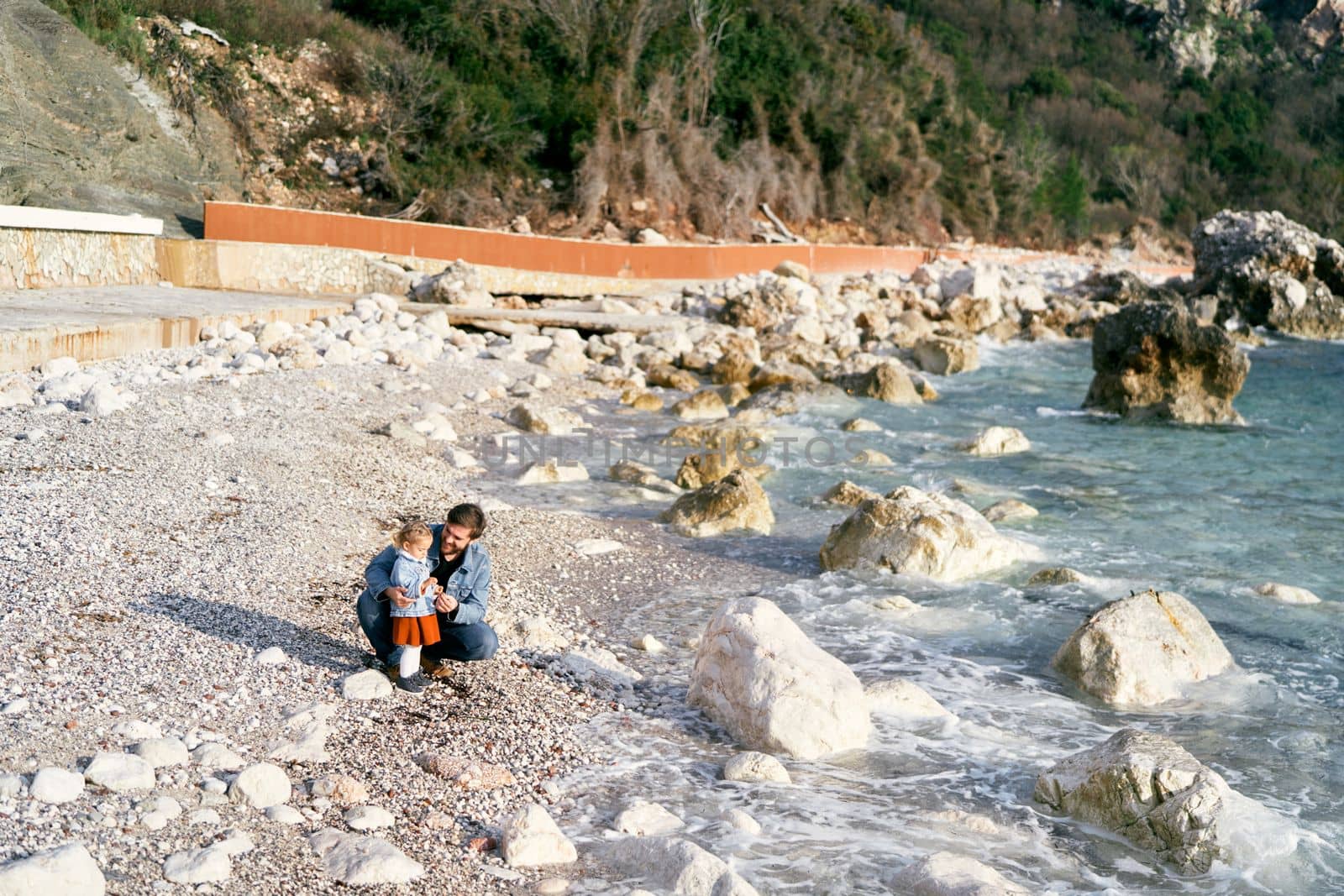 Smiling dad squatted next to a little girl, handing her pebbles from a rocky beach by the water against the backdrop of green mountains. High quality photo