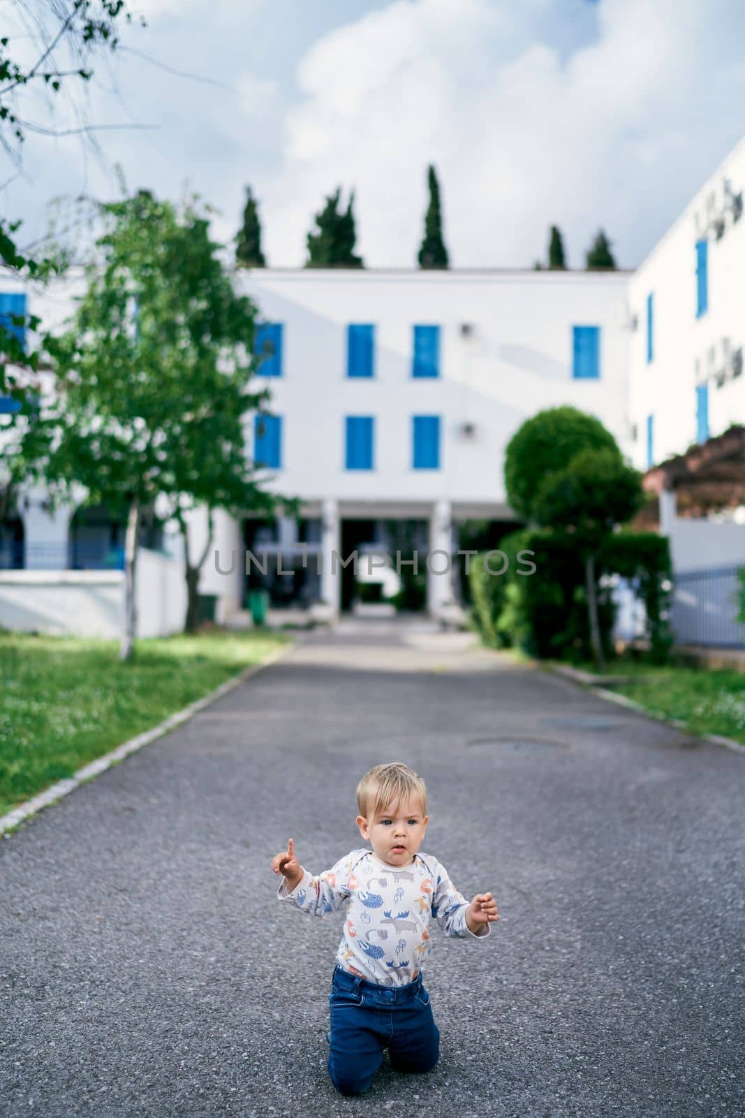 Kid is kneeling on the asphalt against the background of a building and green trees. High quality photo