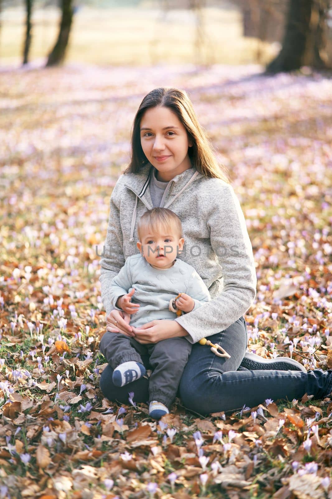 Smiling mother sits on fallen leaves and holds a baby with a rattle in her arms. High quality photo