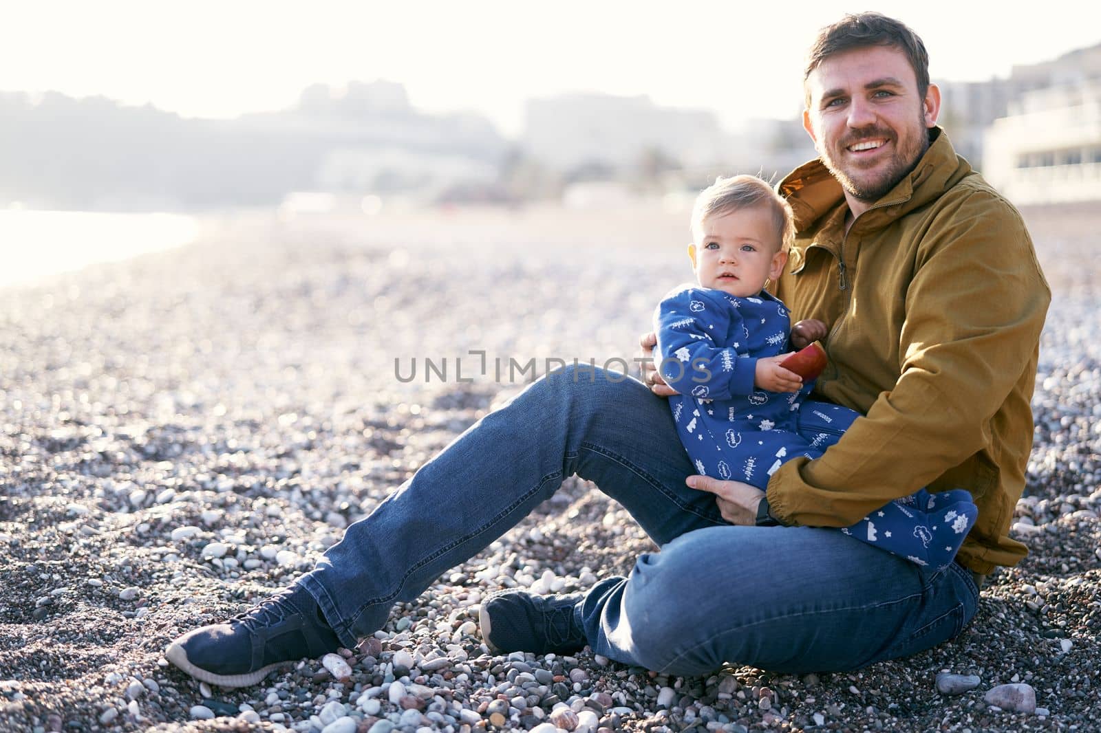 Smiling dad in a brown jacket and jeans holds a little kid in a blue overalls with an apple in his hands while sitting on a pebble beach. High quality photo