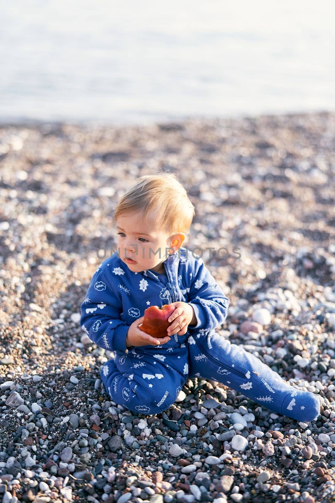 Small baby in a blue overalls sits on a pebble beach and holds an apple. Close-up by Nadtochiy