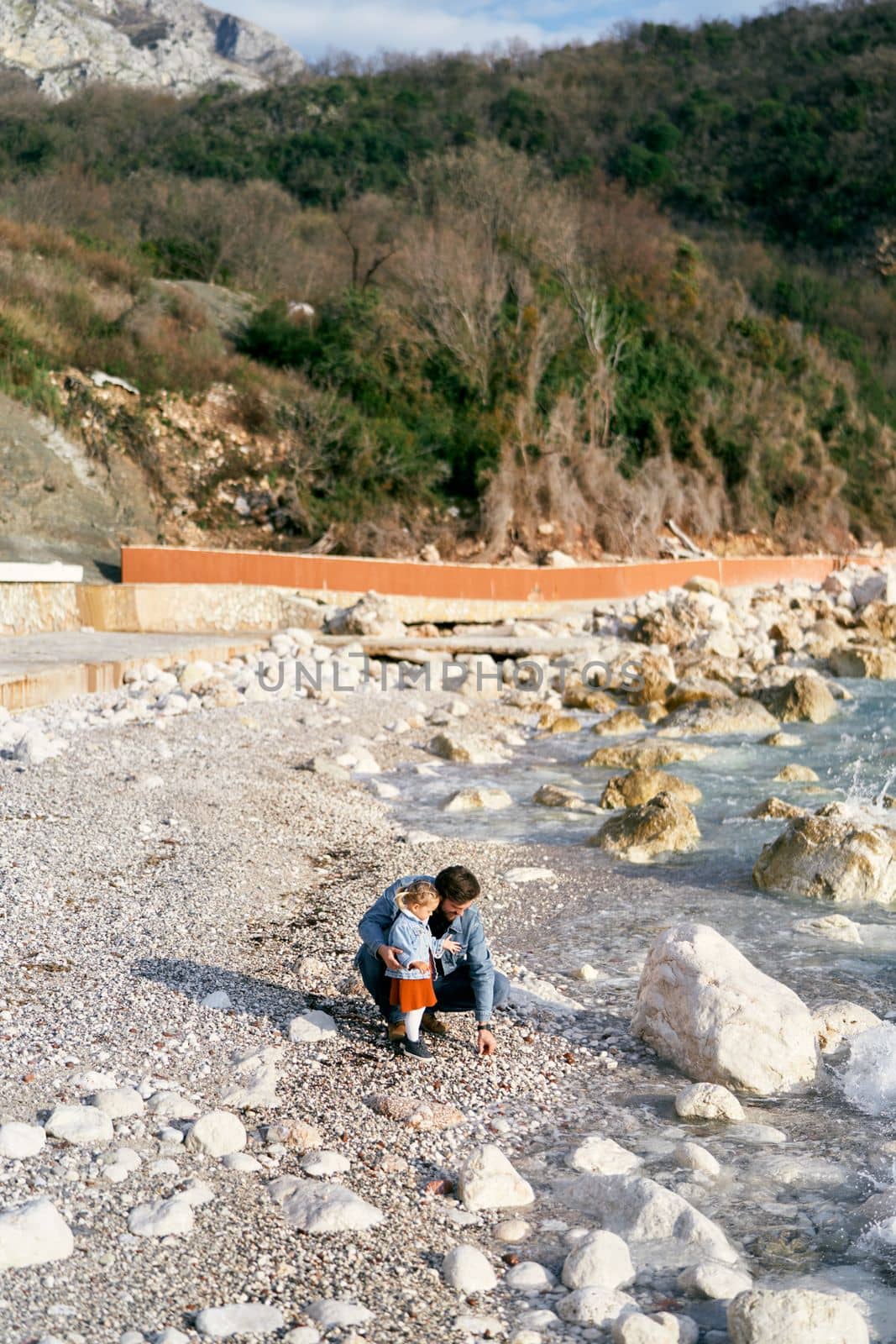 Dad squatted next to a little girl, picking up pebbles from a rocky beach near the water on the background of green mountain. High quality photo