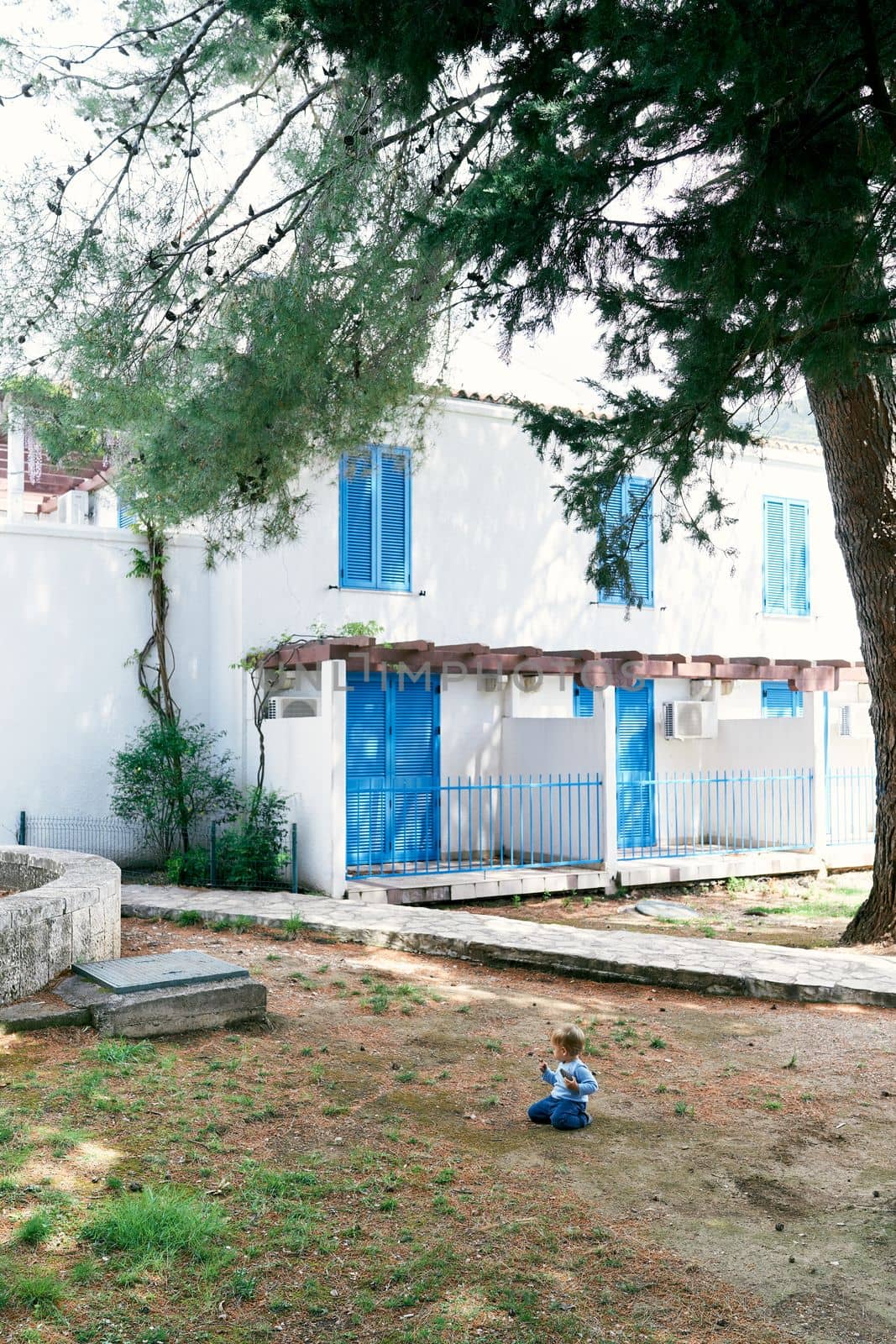 Toddler sits on his knees on the ground in front of a two-story house with closed blinds by Nadtochiy