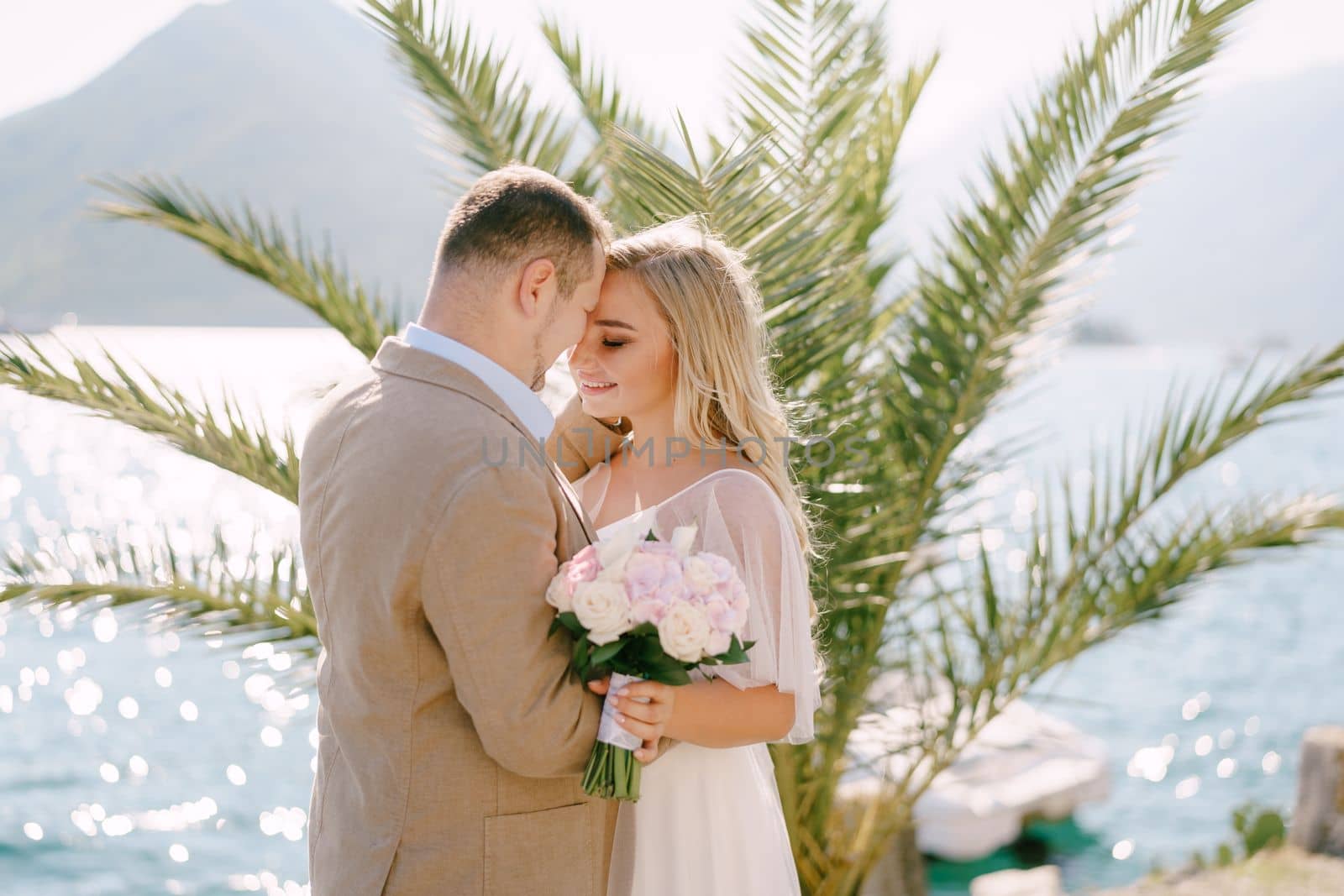 The groom and the smiling bride with a bouquet stand on the pier at the background of a palm tree and tenderly hug . High quality photo