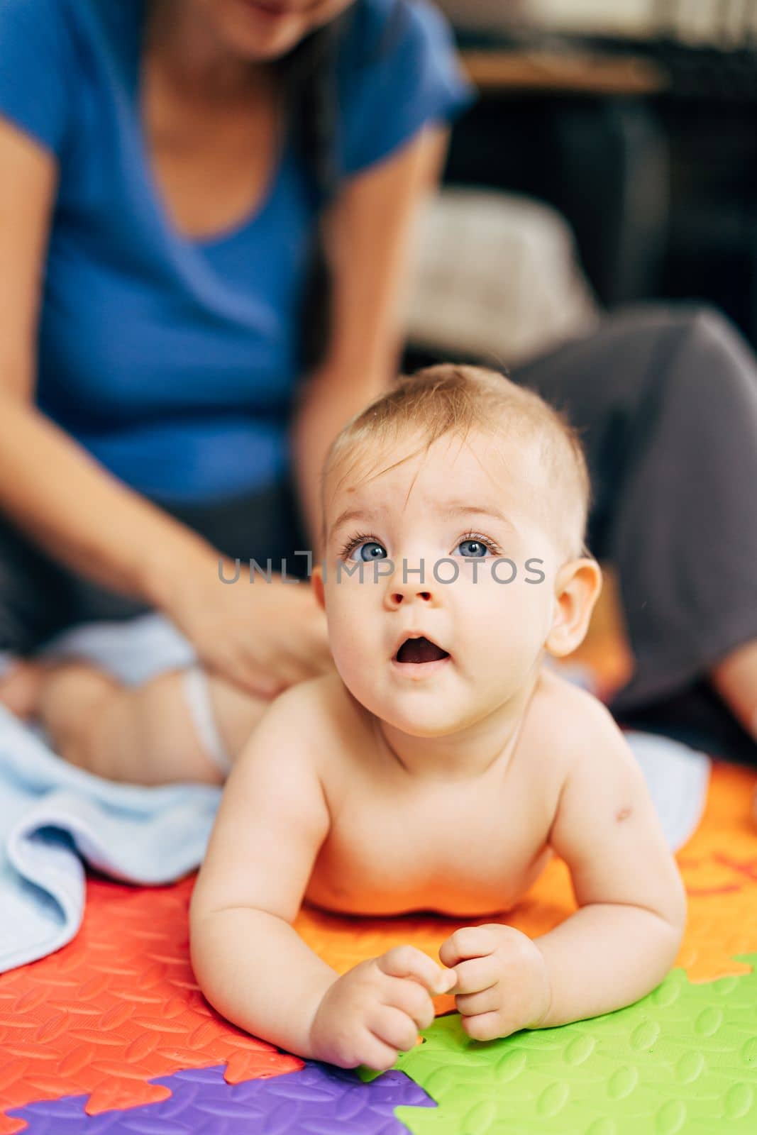 Mom changes diapers to a baby lying on his tummy on a colored rug and pensively looking into the distance by Nadtochiy