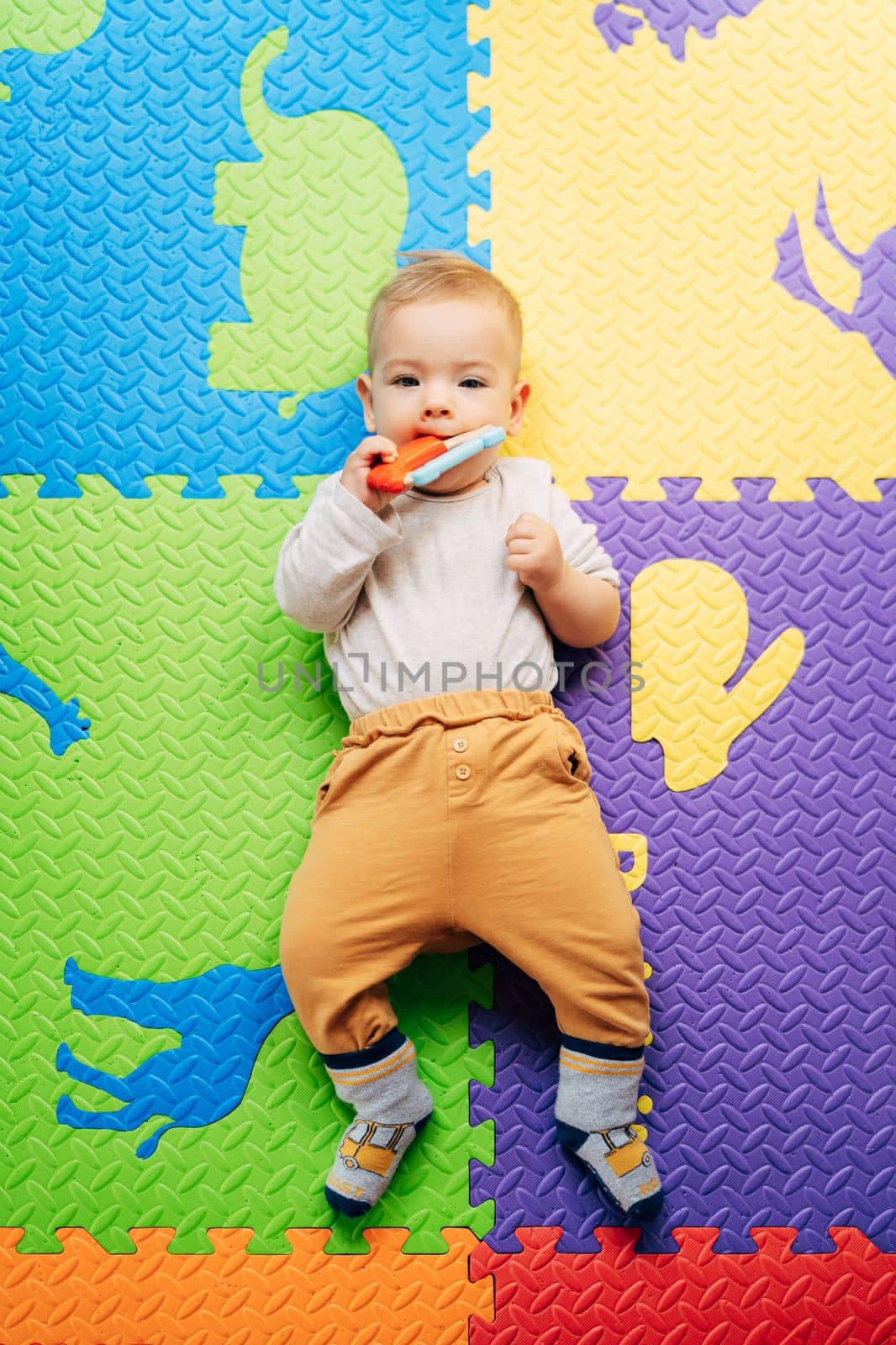 Tiny baby in a white bodysuit and brown pants lies on a colored rug, holds and gnaws on a bright rattle. Top view. High quality photo