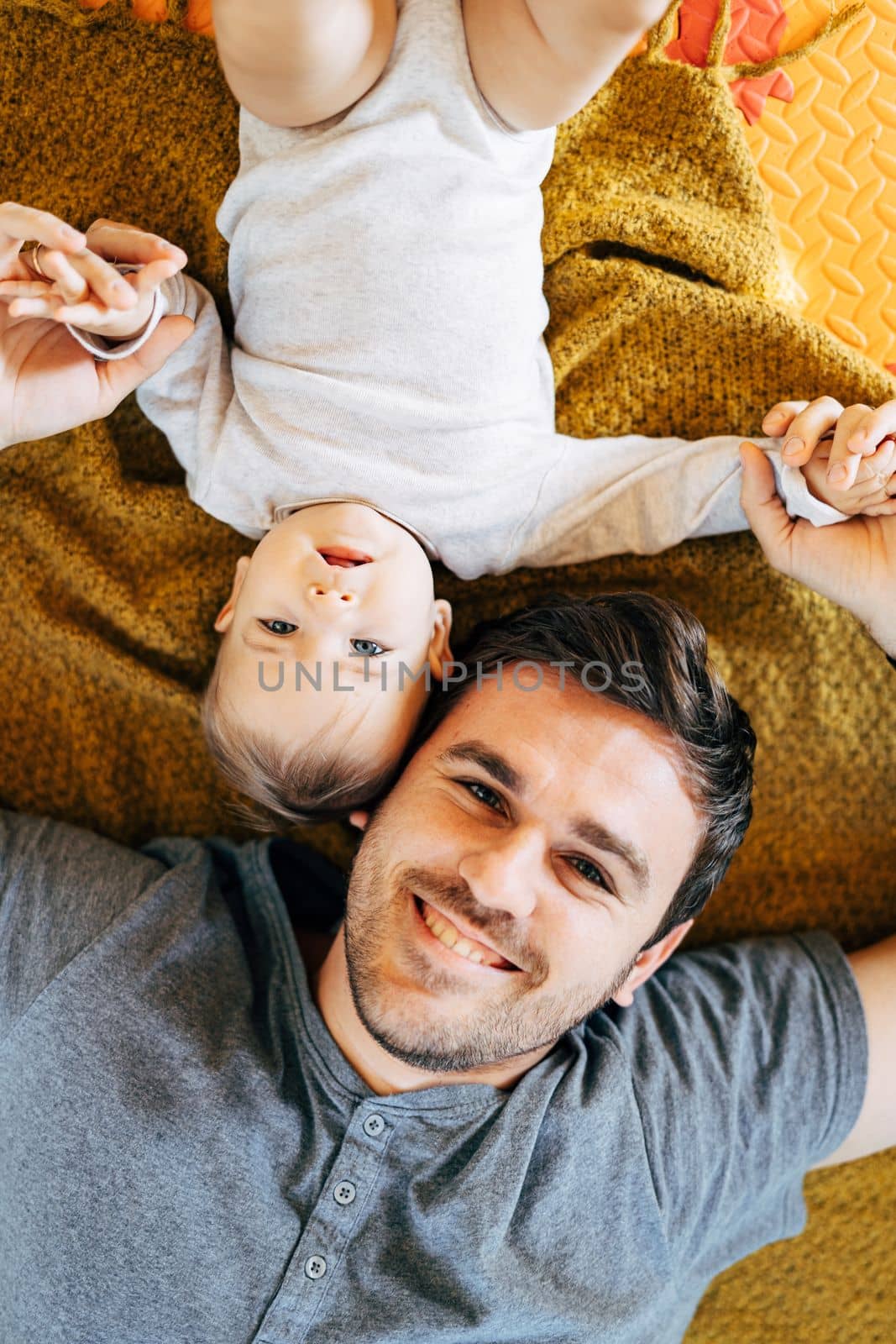 Baby lying on his back head to head smiling dad holding baby's hands on yellow blanket on the floor by Nadtochiy