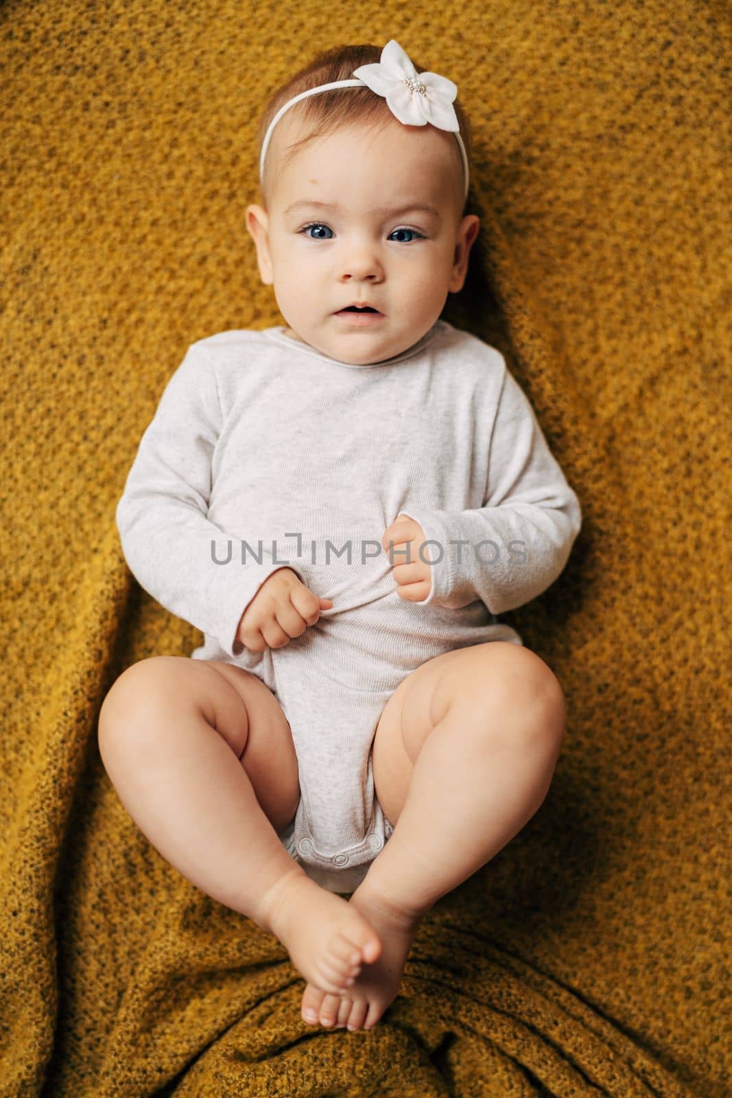 Tiny baby girl with a flower on her head in a white bodysuit lies on a yellow blanket with her hands on her tummy by Nadtochiy