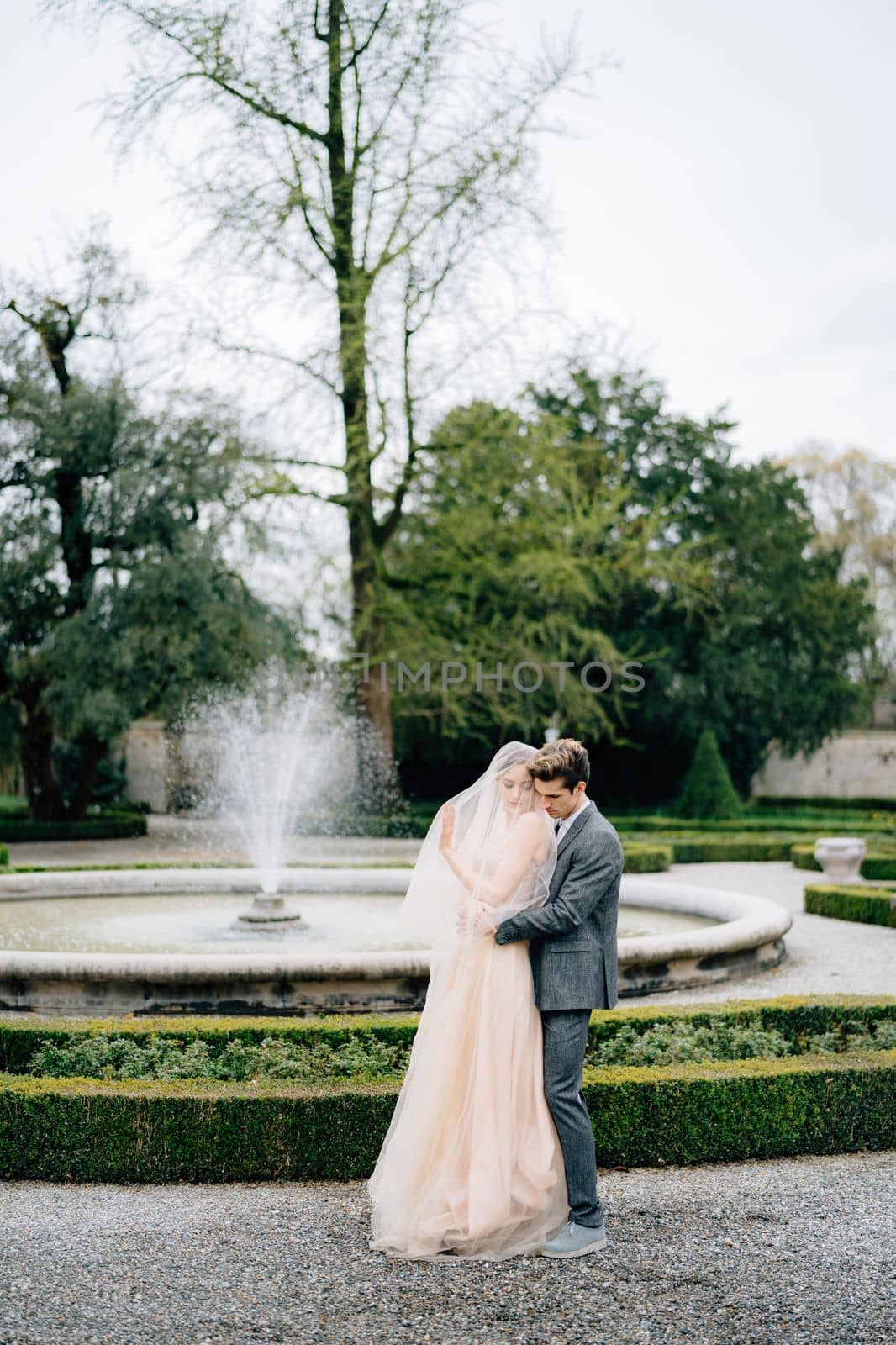 Groom hugs bride in a veil from behind and kisses her on the shoulder, in the garden in front of fountain. Lake Como, Italy. High quality photo