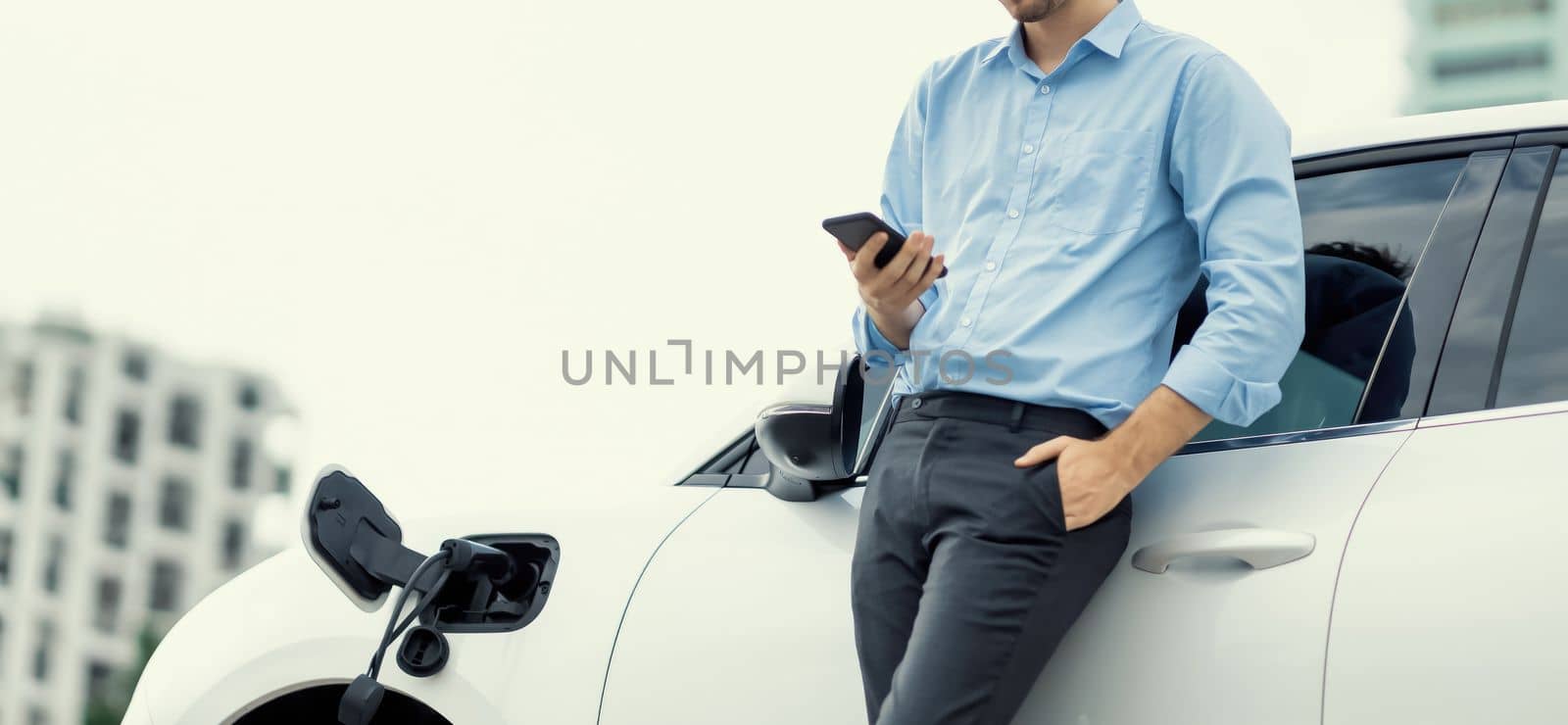 Suit-clad progressive businessman look at EV car's battery status from his phone while standing on a charging station with a power cable plug and a renewable energy-powered electric vehicle.