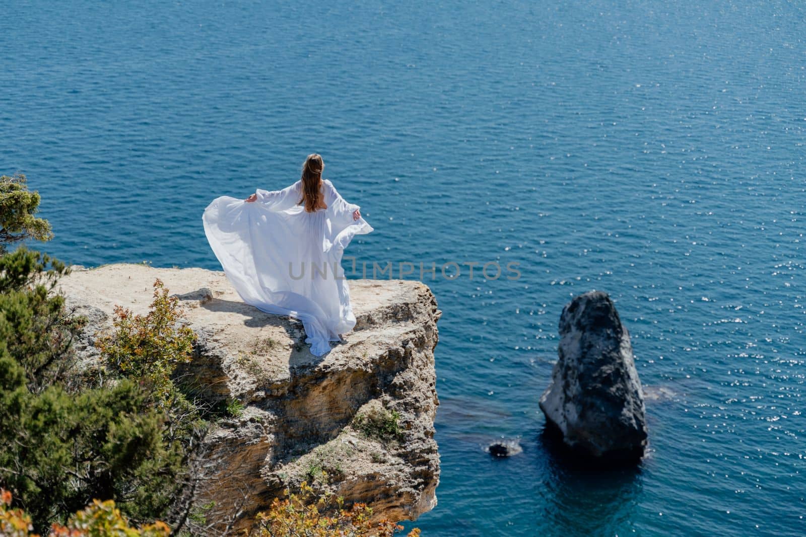 Woman in a white dress on the sea. Side view Young beautiful sensual woman in white long dress posing on a rock high above the sea at sunset. Girl in nature against the blue sky.