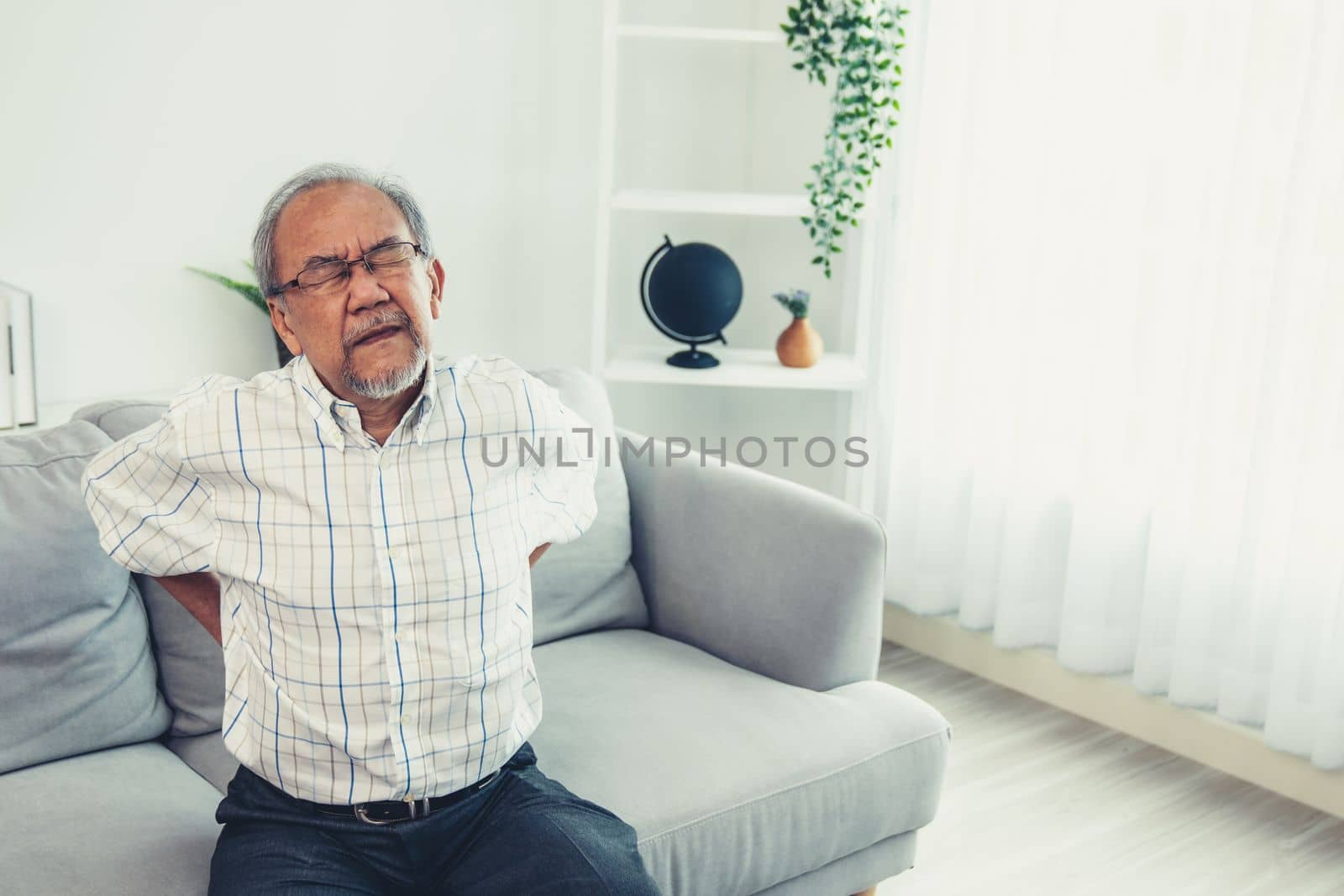 An agonizing senior man in need of assistance while sitting on his sofa at home, suffering from back pain. Senior care, nursing home for pensioners, deteriorating health of old age.