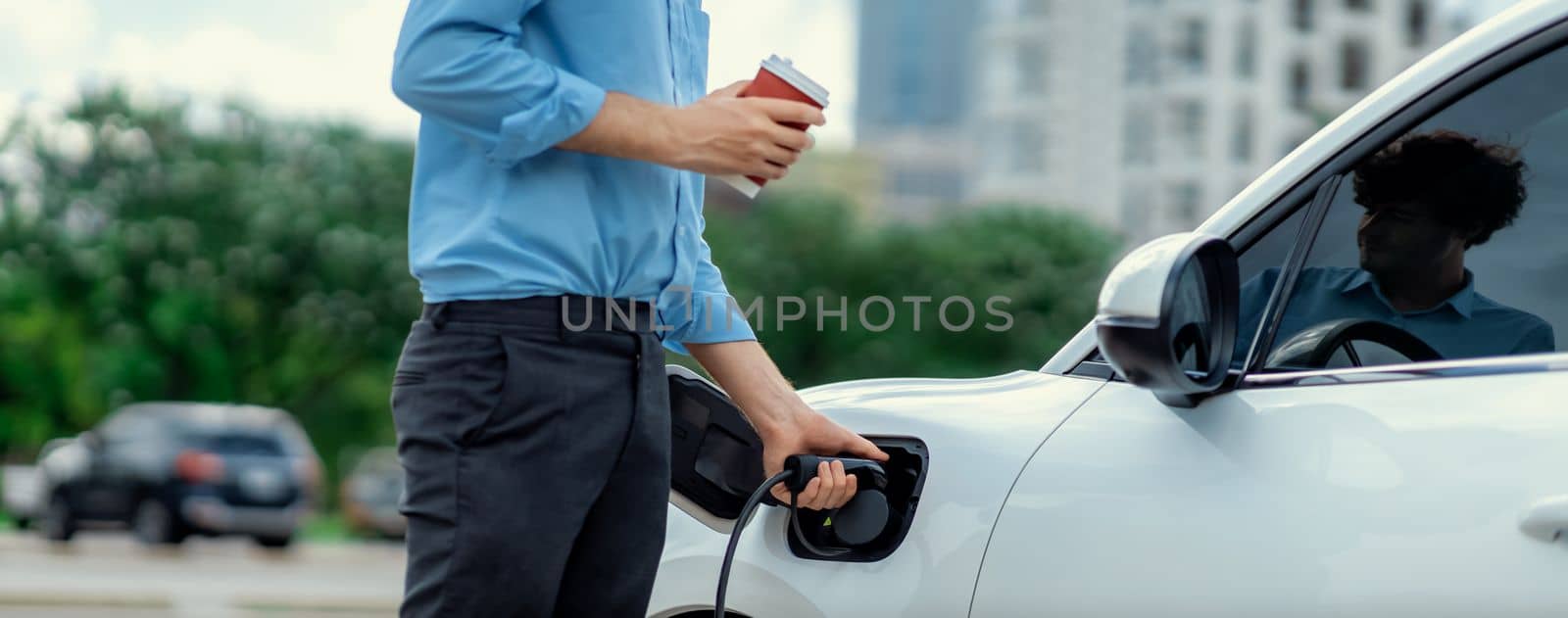 Closeup progressive man holding EV charger plug from public charging station for electric vehicle with background of residential building as concept eco-friendly sustainability energy car concept.