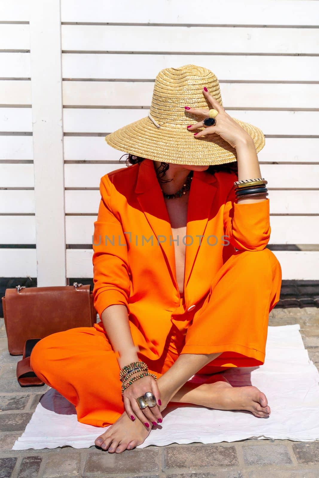 Stylish woman in an orange suit with a hat sits on a rug on a white striped background. On the hands are jewelry rings and bracelets, sandals and a bag stand side by side