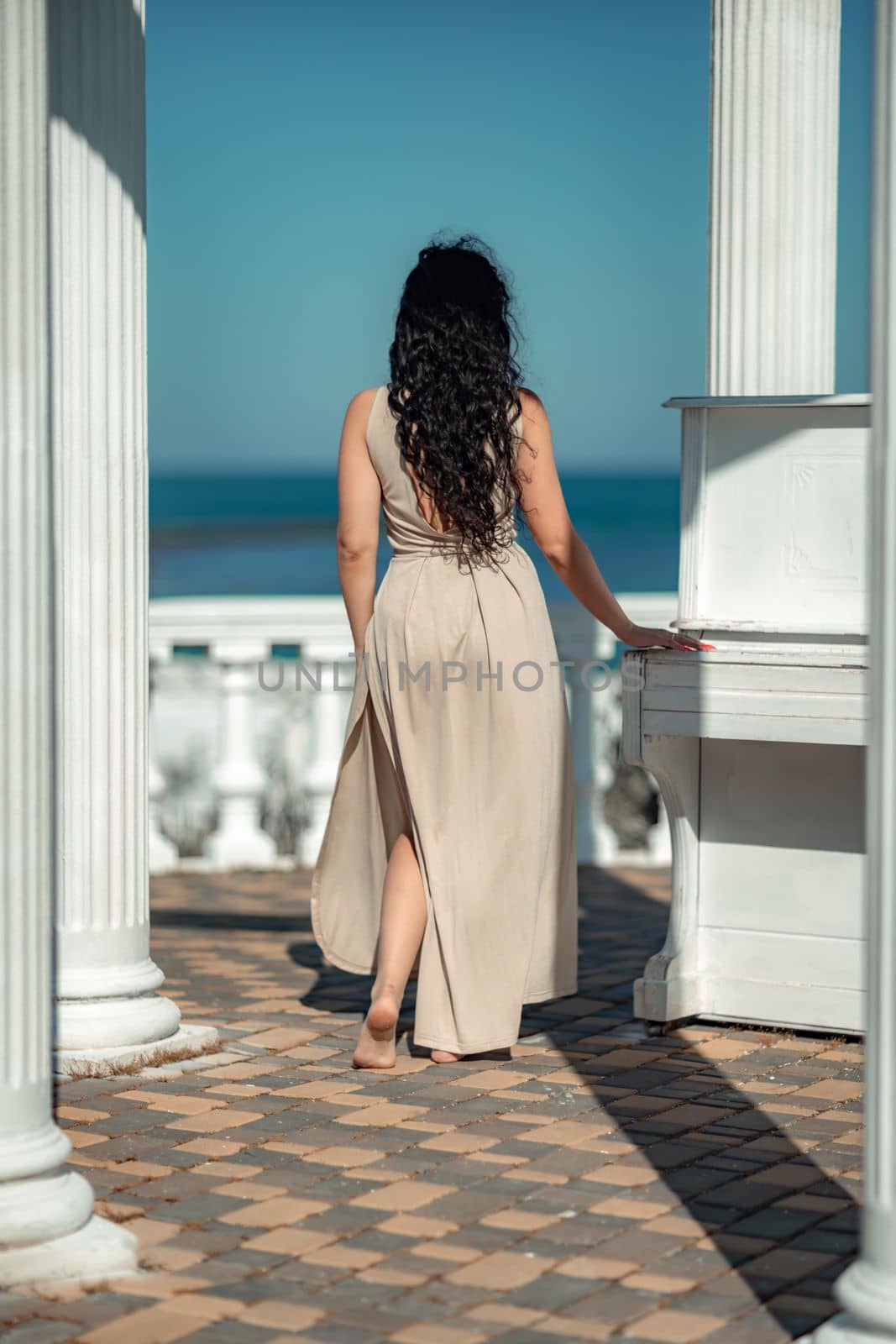 Sea woman rest. A woman with long curly hair in a beige dress stands with her back and looks at the sea and the coast from a balcony with balusters. Tourist trip to the sea. by Matiunina