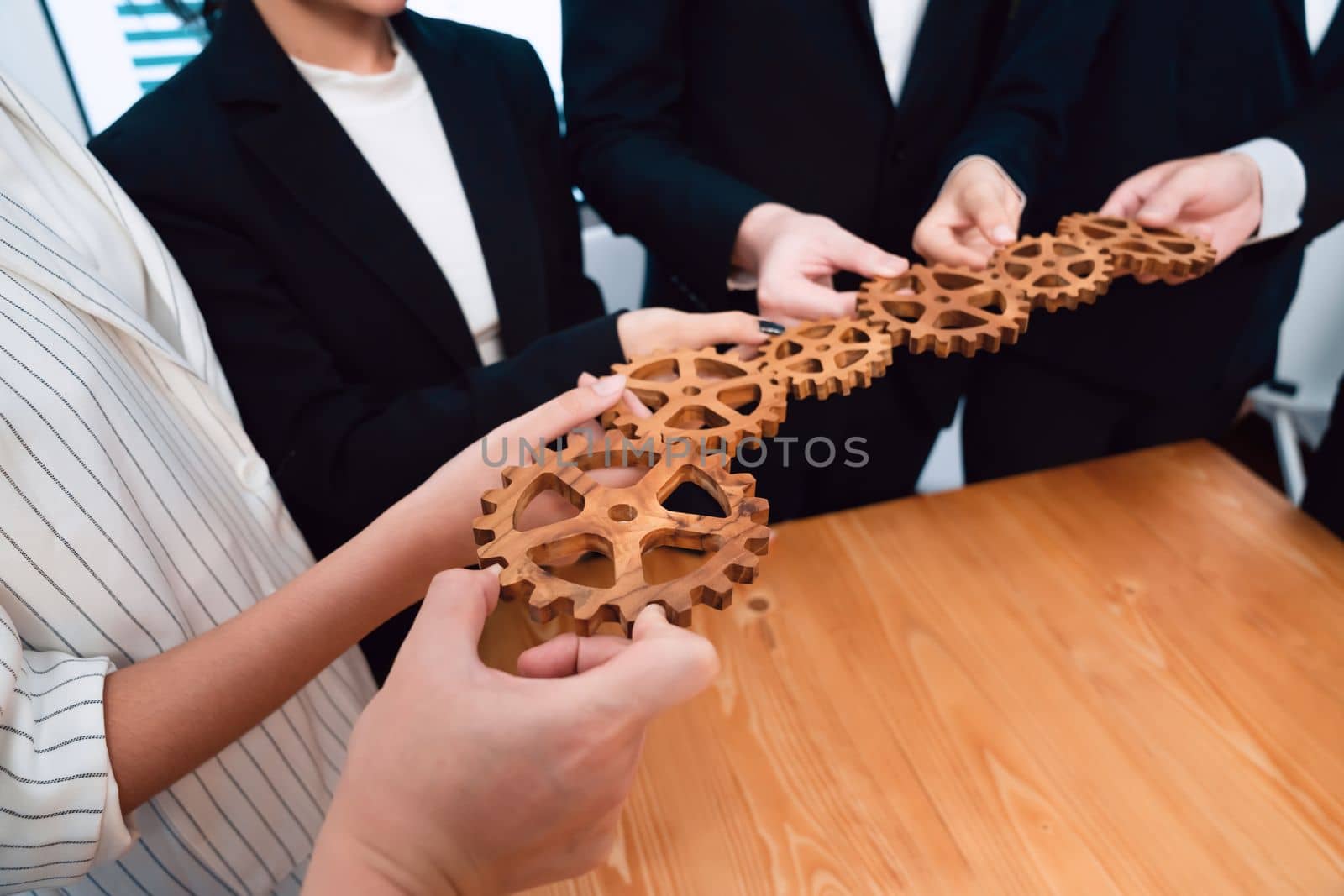 Closeup hand holding wooden gear by businesspeople wearing suit for harmony synergy in office workplace concept. Group of people hand making chain of gears into collective form for unity symbol.